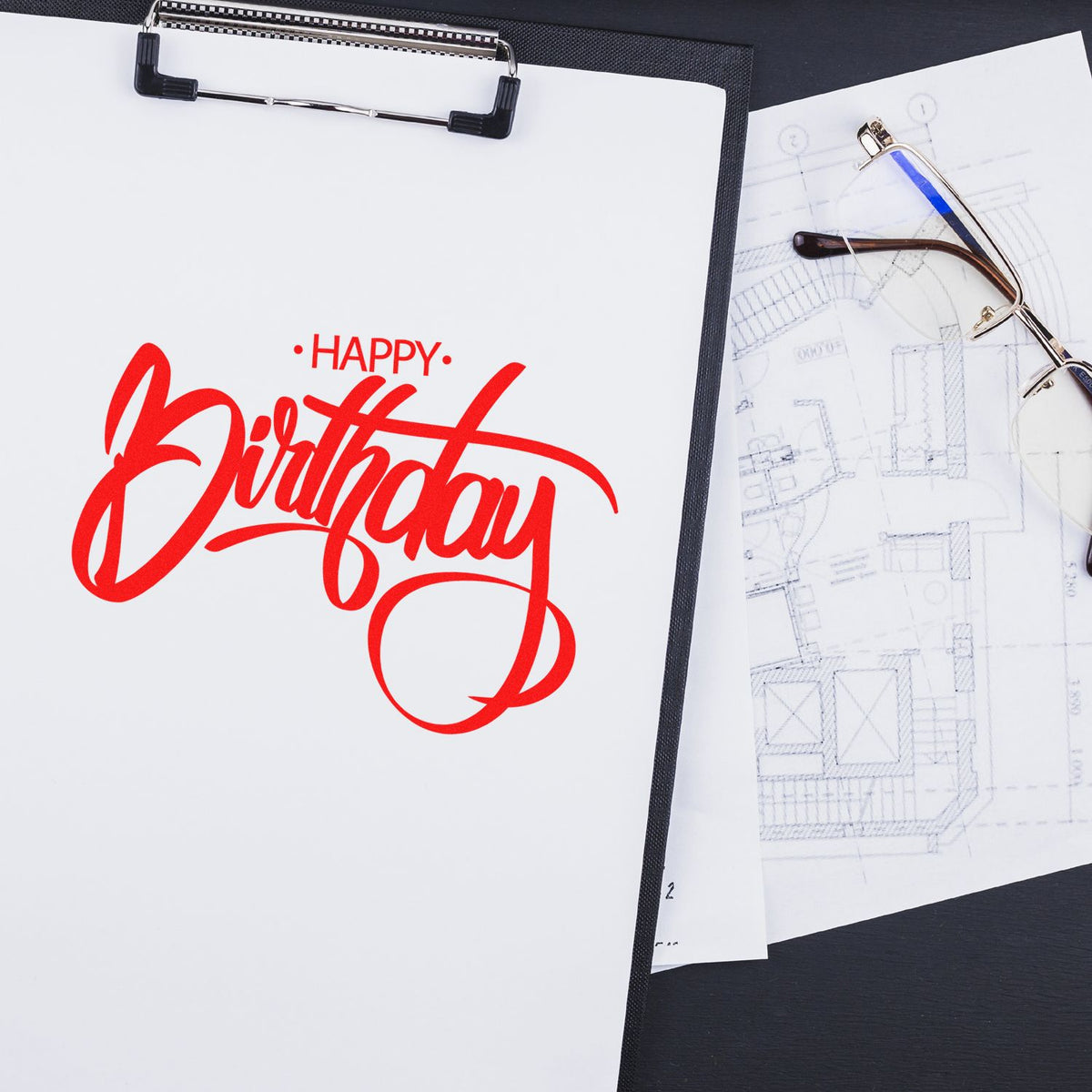 Red Happy Birthday stamp on white clipboard beside eyeglasses and blueprint, showcasing stamp and seal products.