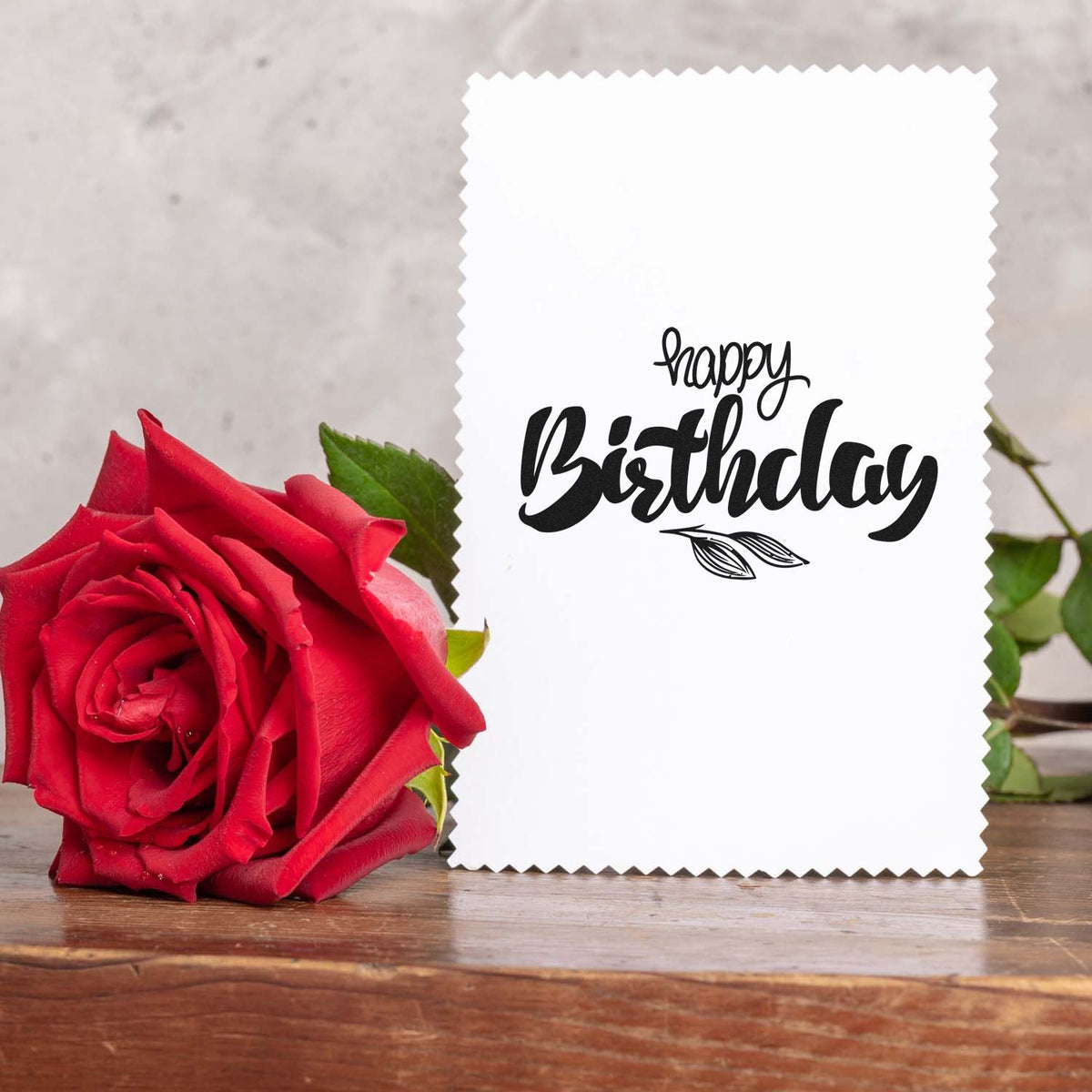White scalloped birthday stamp seal card with black lettering, beside a red rose on a rustic wooden table.