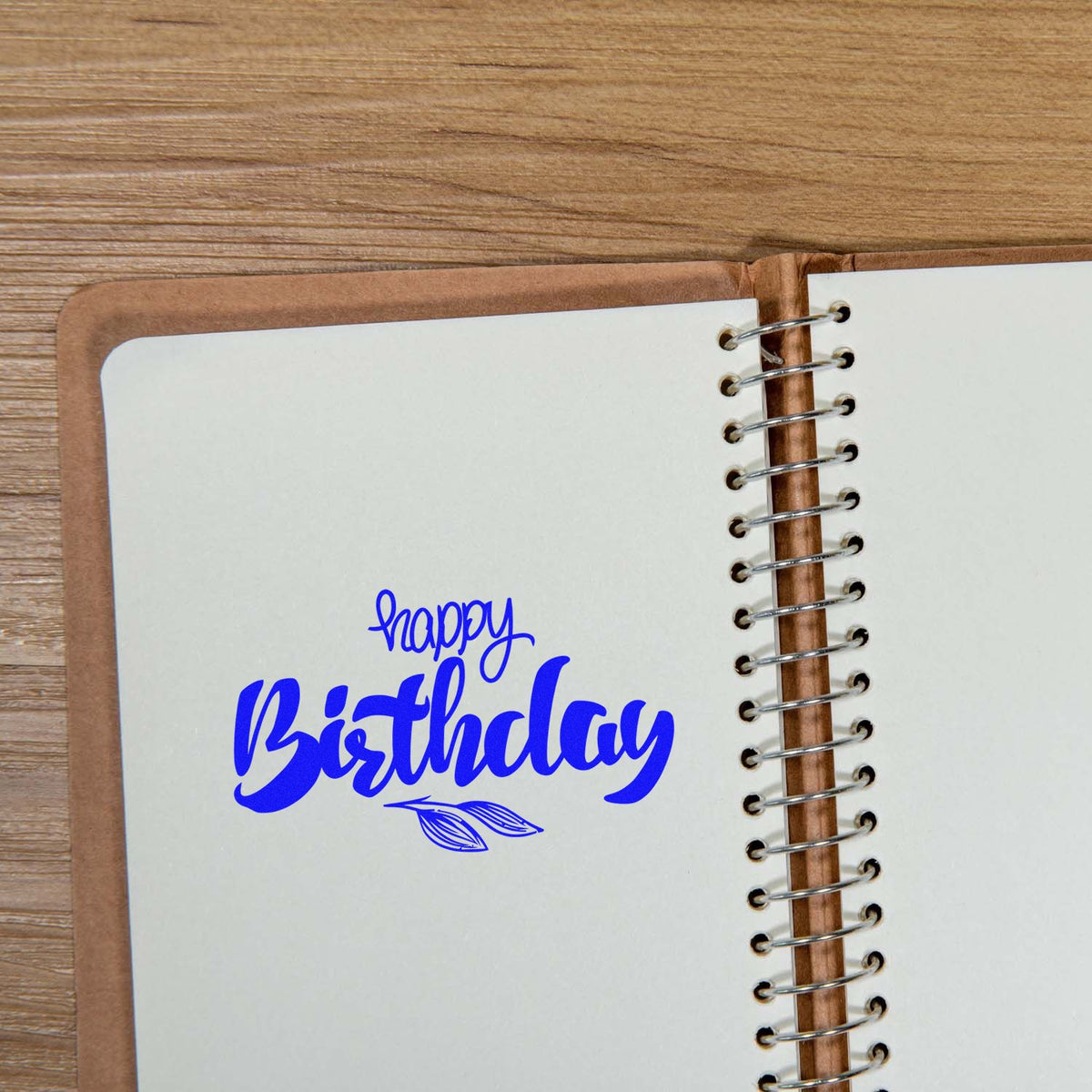 Blue happy Birthday stamp and leaf seal on a white spiral notebook page, on a wood table.