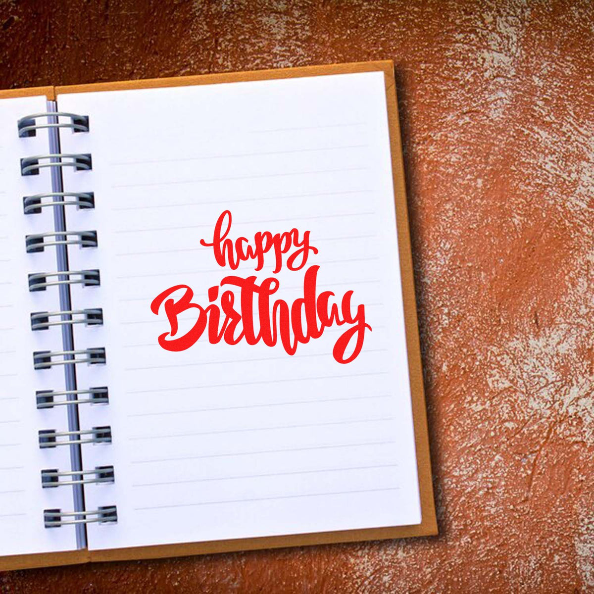 Red Happy Birthday stamp on white spiral notebook with silver rings, set against a textured brown background.