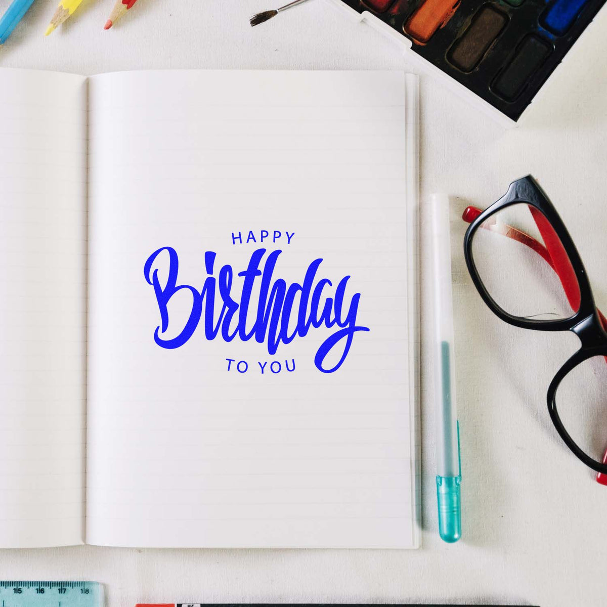 Blue Happy Birthday stamp and seal on an open notebook, with watercolor palette, glasses, and pen on a white desk.