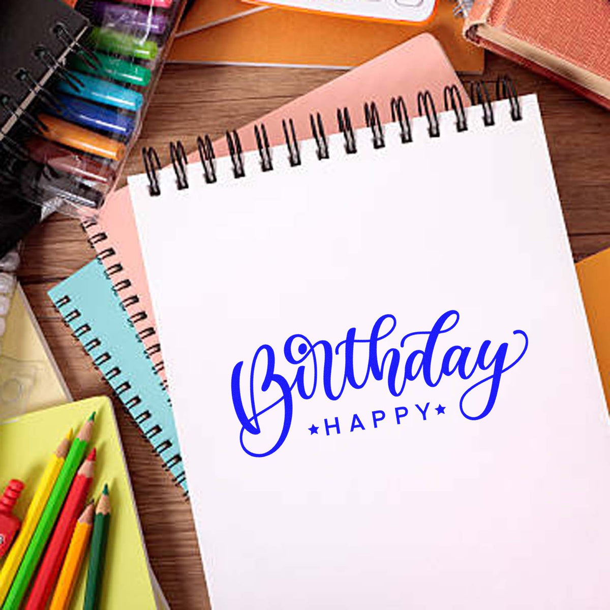 Blue Birthday Happy stamp/seal design on a white spiral notebook, with colorful notebooks and pencils on a wooden desk.