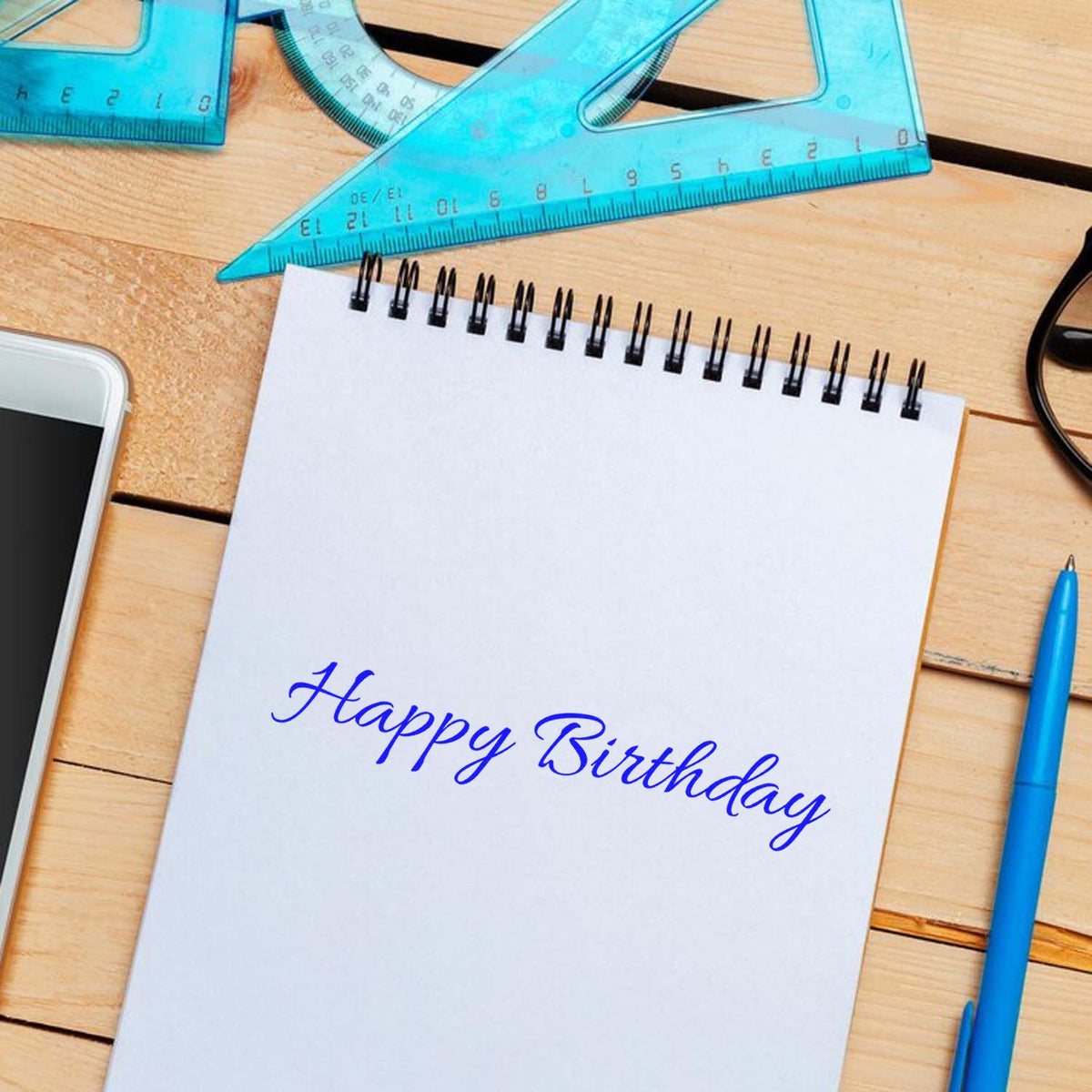 Blue Happy Birthday stamp/seal on a white spiral notebook, with turquoise rulers, blue pen, and wooden desk.