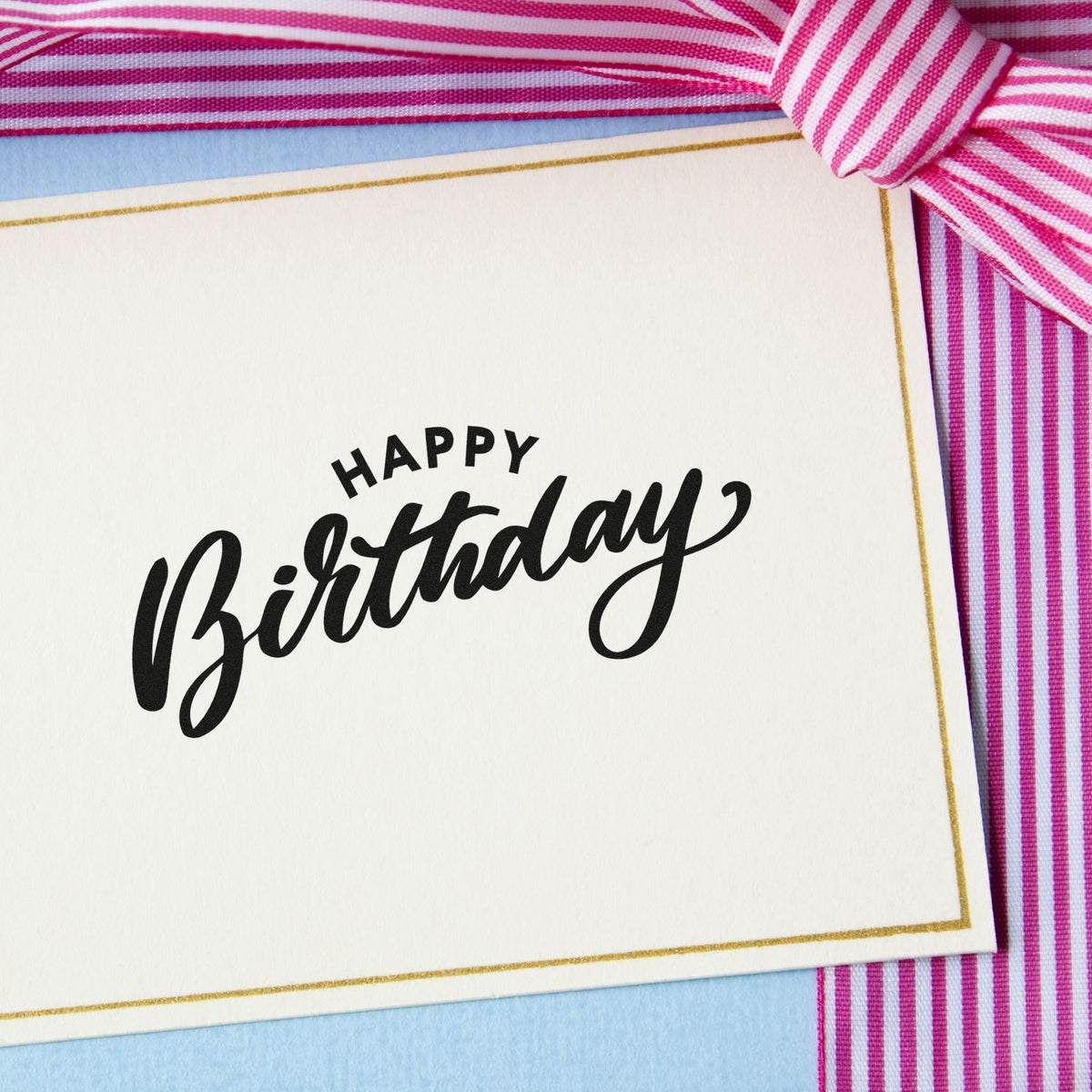 Black Happy Birthday stamp seal on white card with gold border, pink striped ribbon, and light blue background.