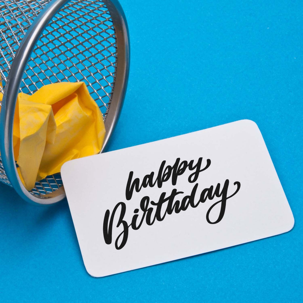 White birthday stamp and seal card with black script, beside a silver mesh basket, yellow tissue, and bright blue background.