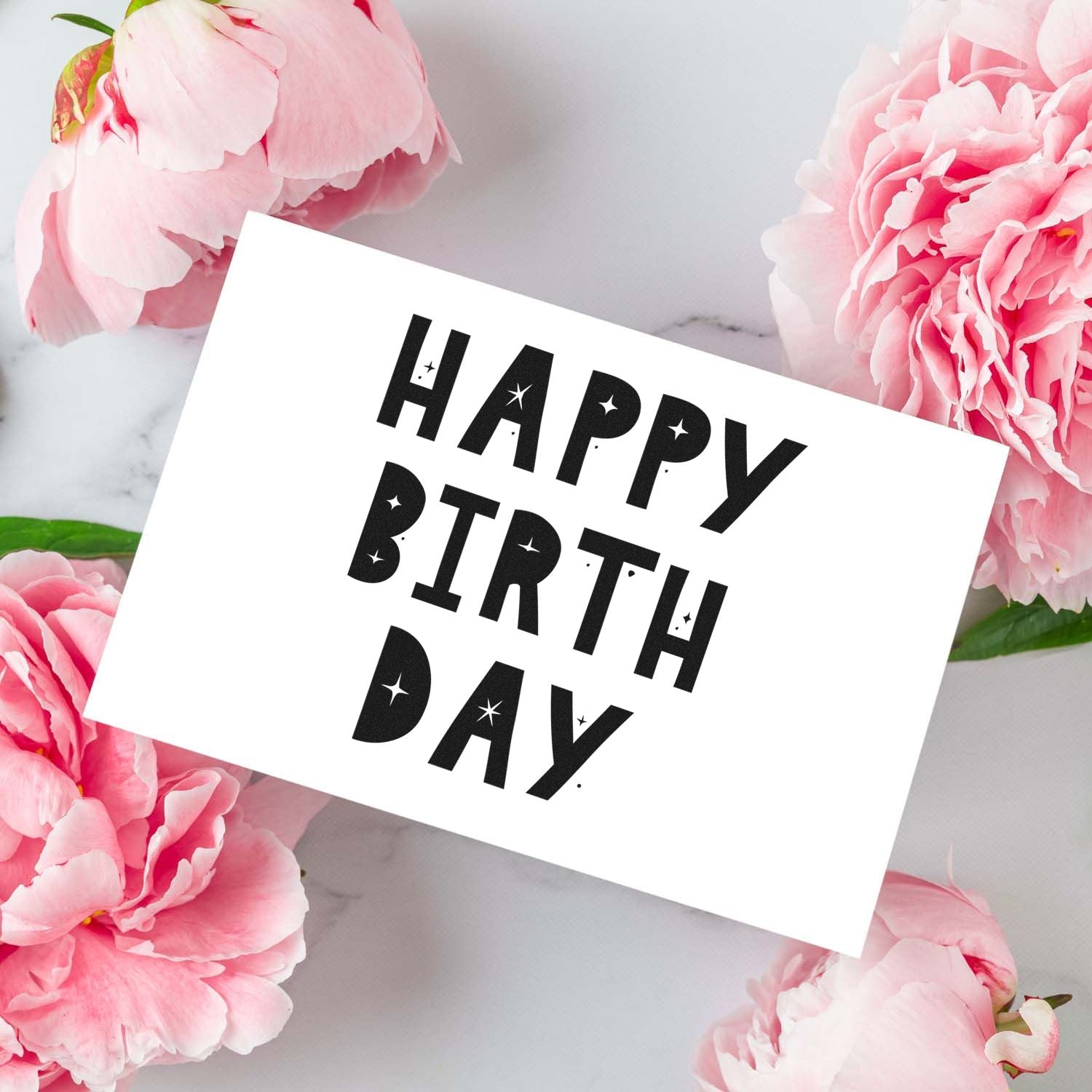 Happy Birthday black stamp and seal on a white card, framed by pink peonies on a marble background.