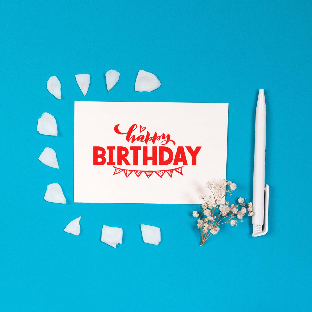 White birthday stamp seal with red lettering on bright blue background, surrounded by white petals and a white pen.