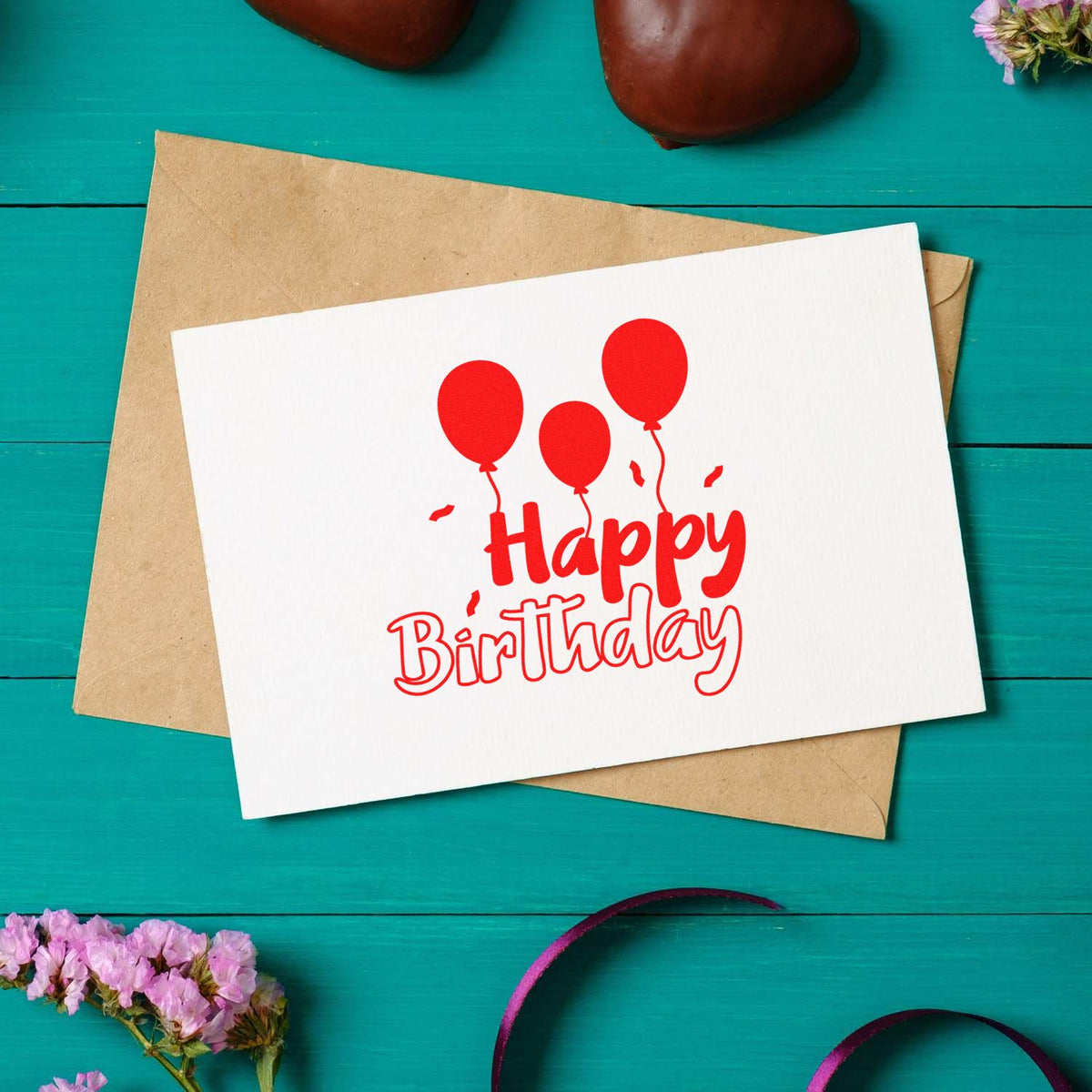 Red Happy Birthday stamp with balloon seal on white card over kraft envelope, on teal wooden background.
