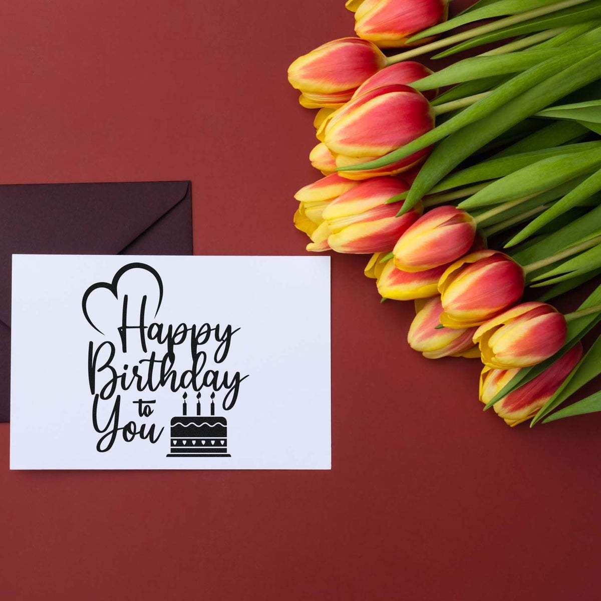 Birthday stamp and seal on white card with black script and cake icon, beside pink-yellow tulips on burgundy background