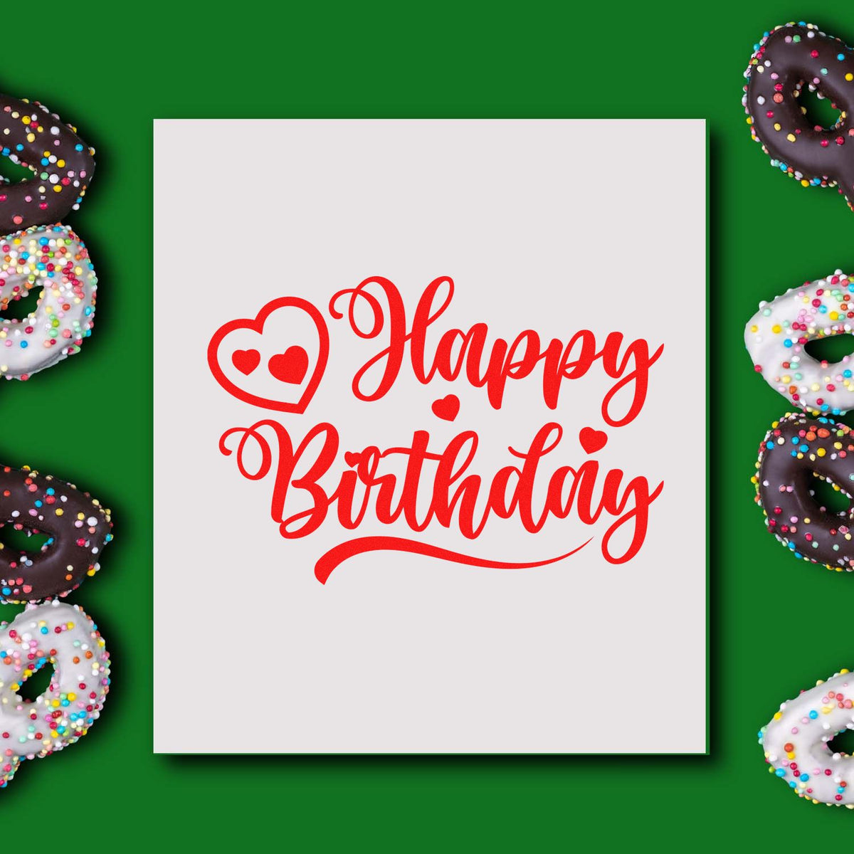Red Happy Birthday stamp and seal on white card, surrounded by sprinkle-covered chocolate and white donuts on green background.