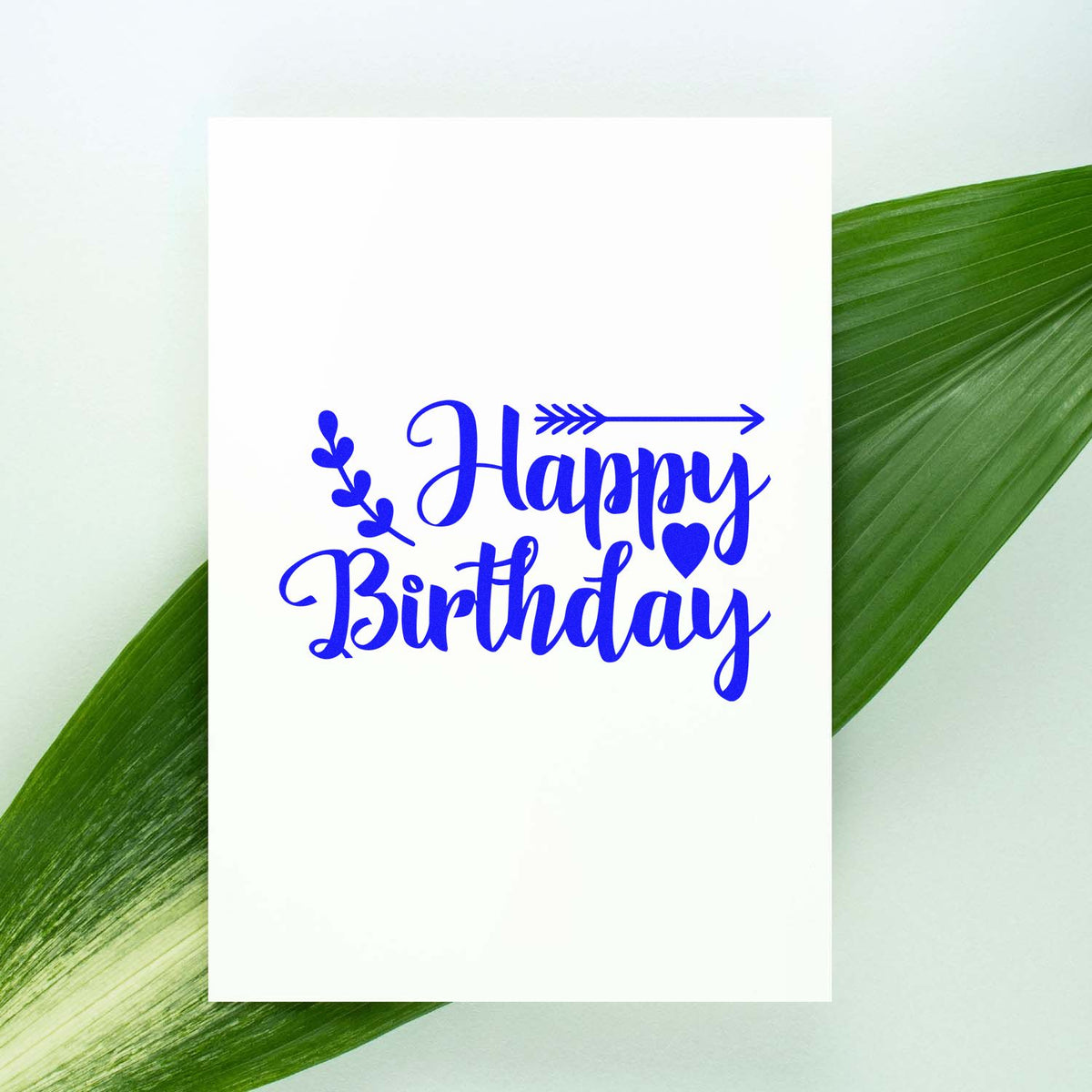 Blue Happy Birthday stamp and seal design with heart and arrow on white card, framed by green leaves.