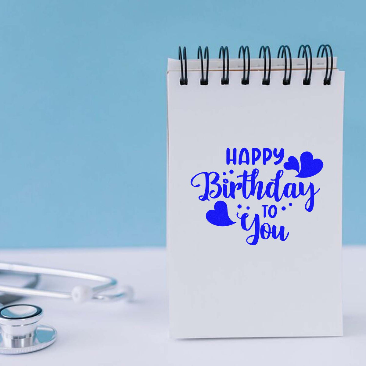 Blue Happy Birthday to You stamp and heart seal on a white spiral notebook against a light blue background.