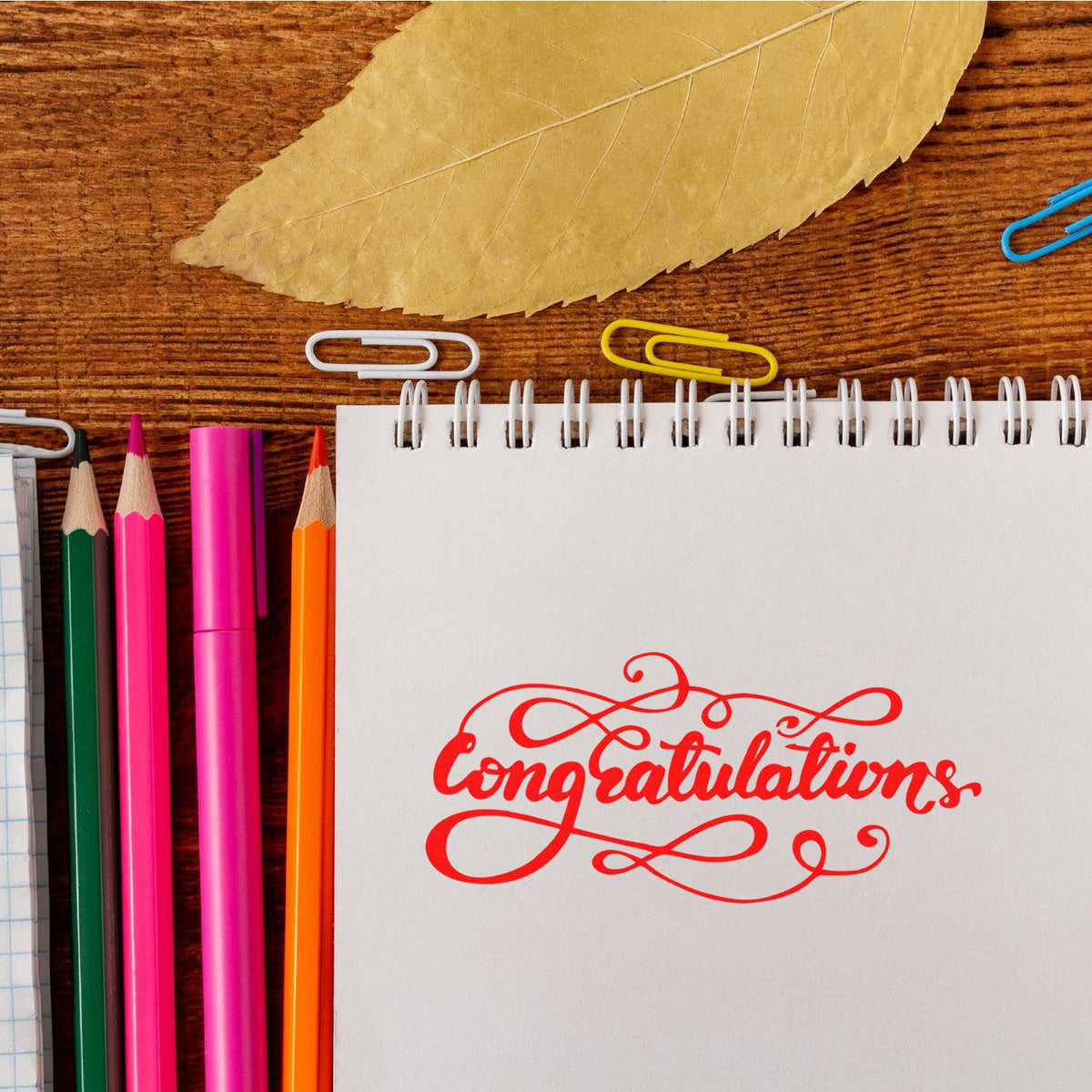 Red congratulations stamp on white spiral notebook, with colorful pencils, paper clips, and a beige leaf on wood desk.