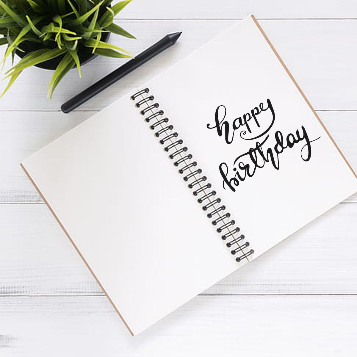 Black happy birthday stamp seal on a white spiral notebook, with a black pen and green plant on a white wood desk.