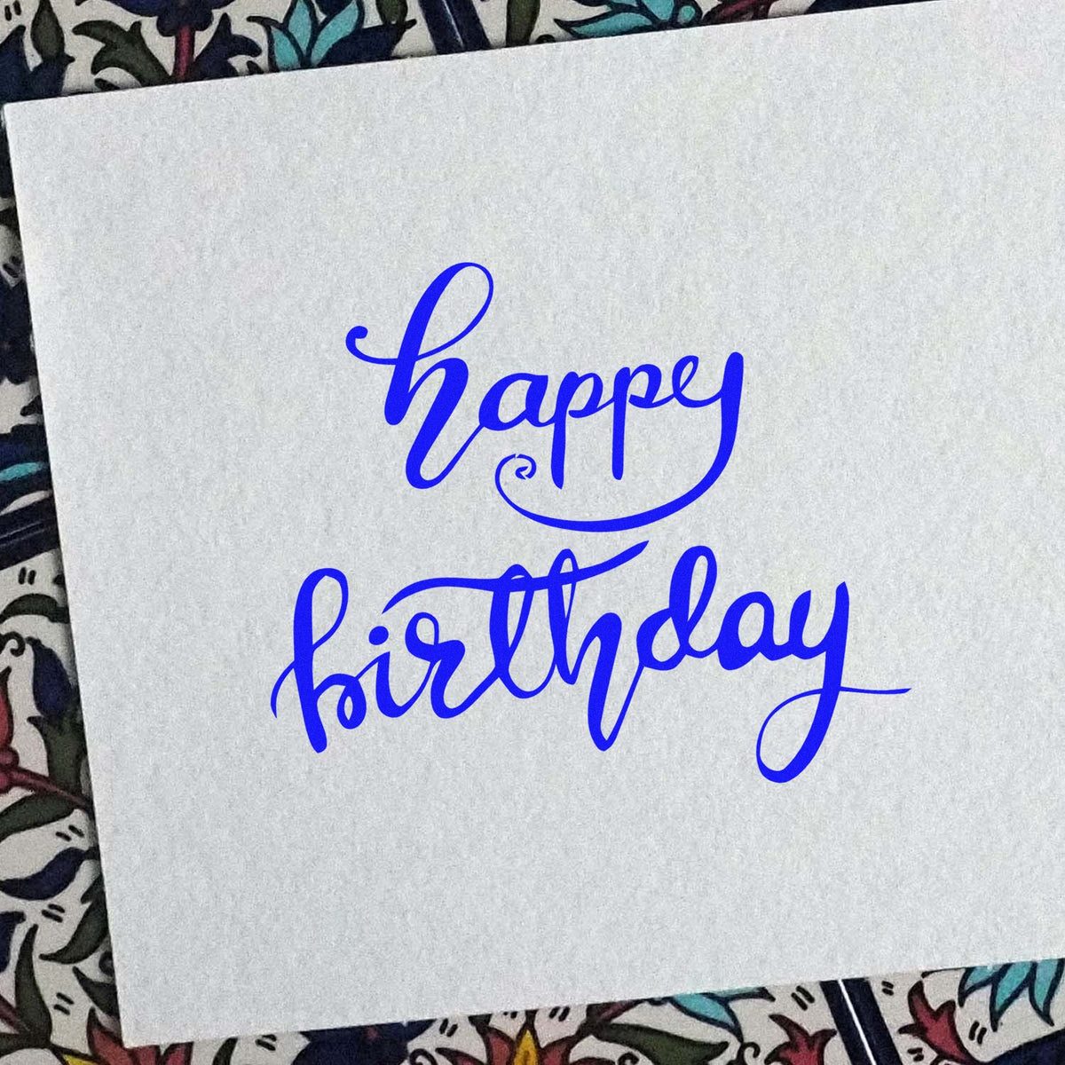 Blue calligraphy Happy Birthday stamp on white paper seal, with decorative floral background.
