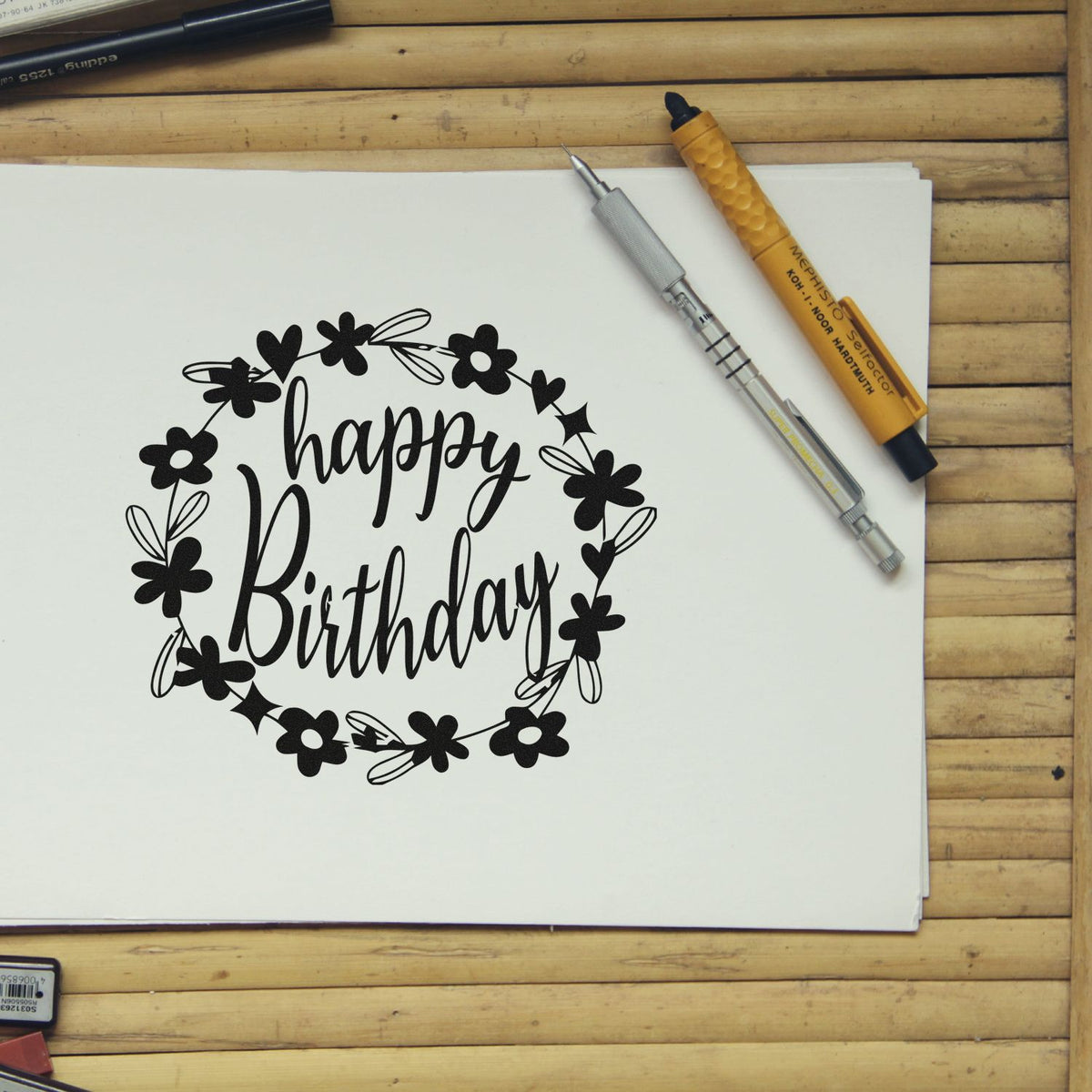 Black happy birthday stamp with floral wreath on white paper, plus silver and yellow seal pens on a wooden table.
