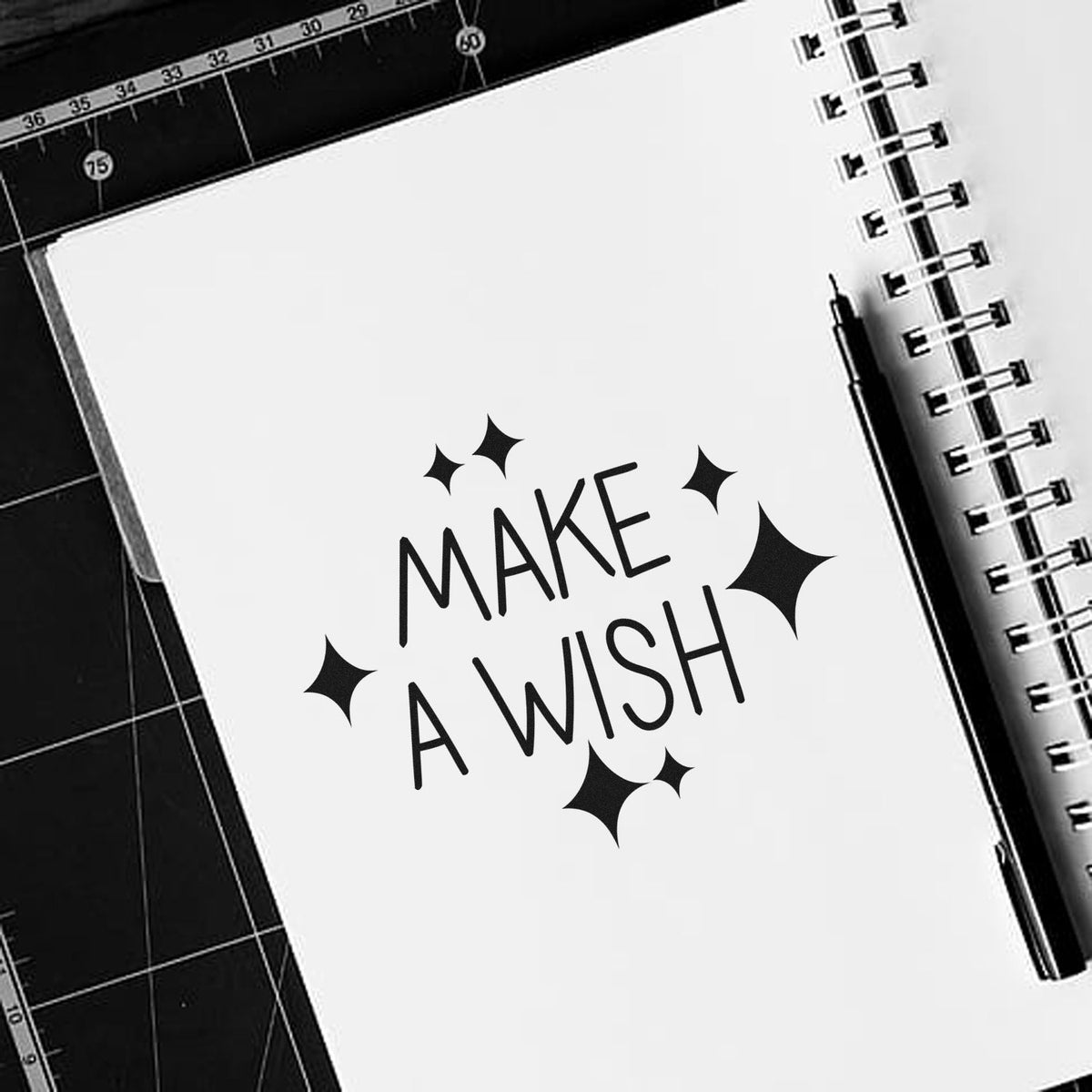 Black MAKE A WISH stamp with sparkles on a white notebook page, beside a spiral binding and pen on a black cutting mat.