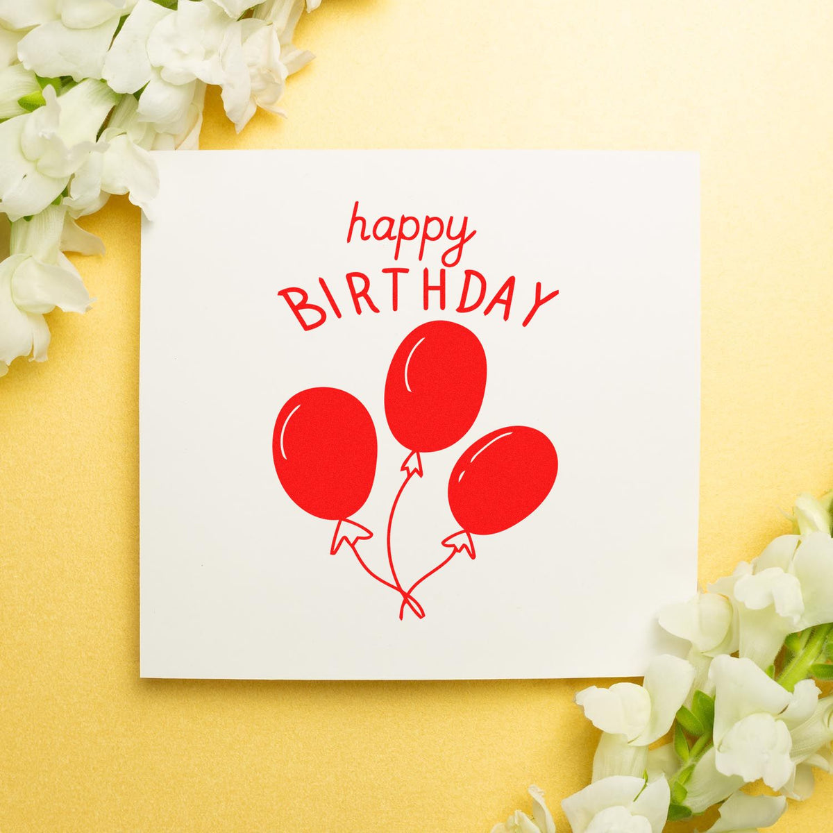 Red happy birthday stamp and seal with balloon design on white card, surrounded by white flowers on pale yellow background.