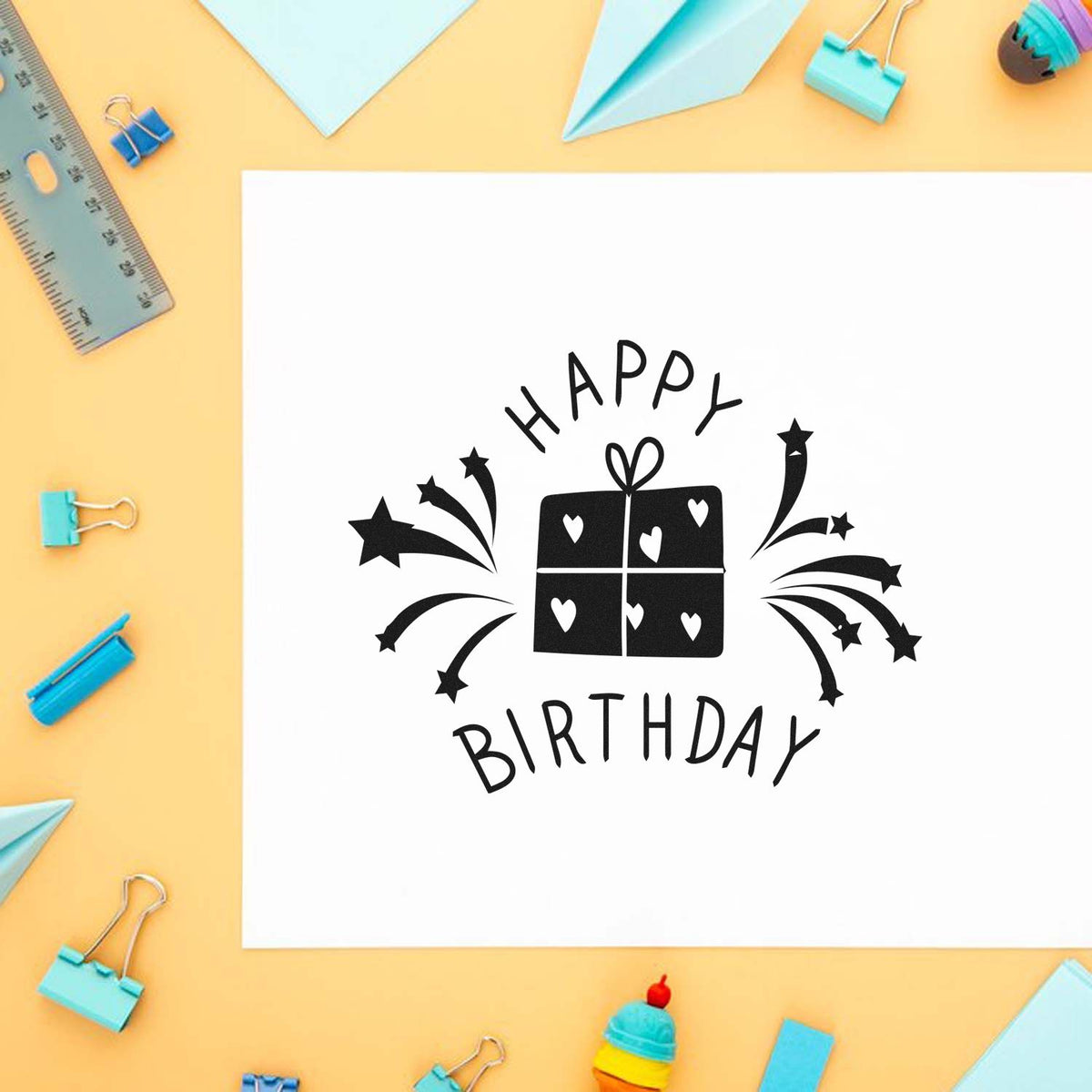 Black Happy Birthday stamp and seal with gift icon on white paper, surrounded by pastel blue stationery on a peach background.
