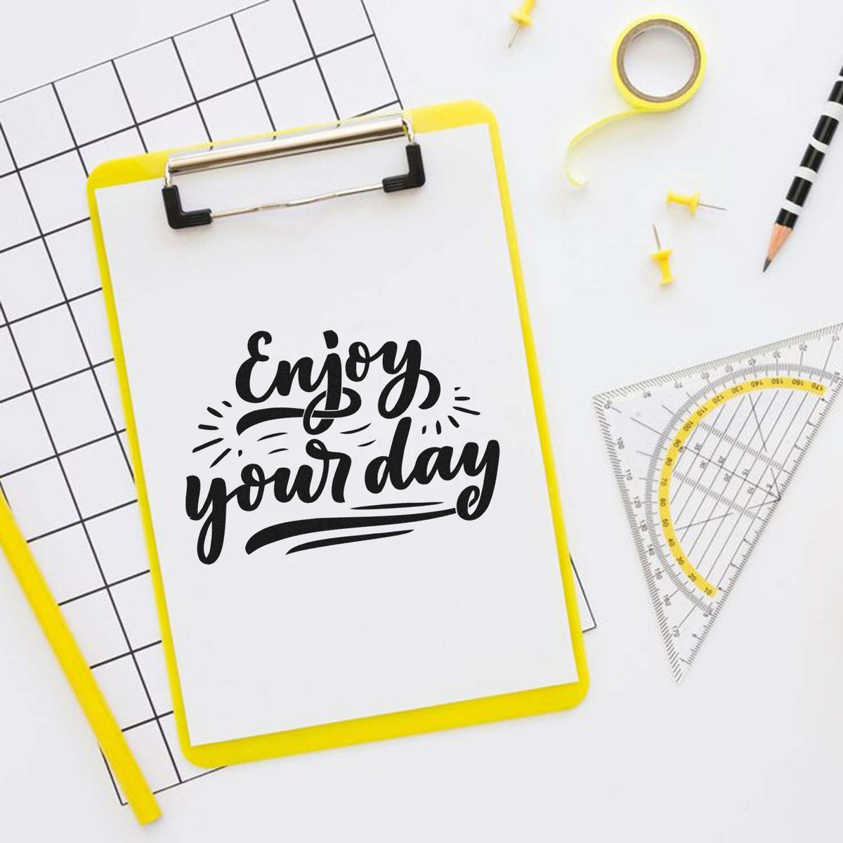 Yellow stamp and seal clipboard with black Enjoy your day lettering, grid paper, ruler, tape, and pencil on white background.