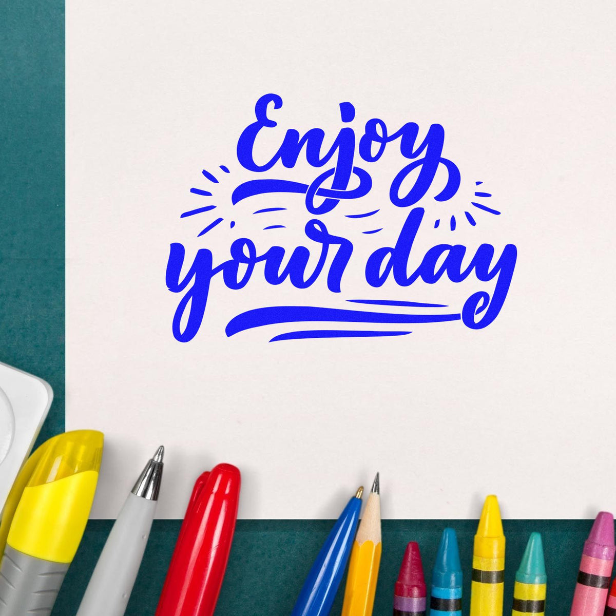 Blue Enjoy your day stamp seal on white paper, surrounded by colorful pens and markers.