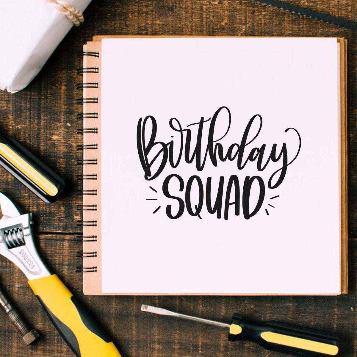 Birthday Squad black script stamp on white spiral notebook, with yellow-and-black seal tools on a rustic wood desk.
