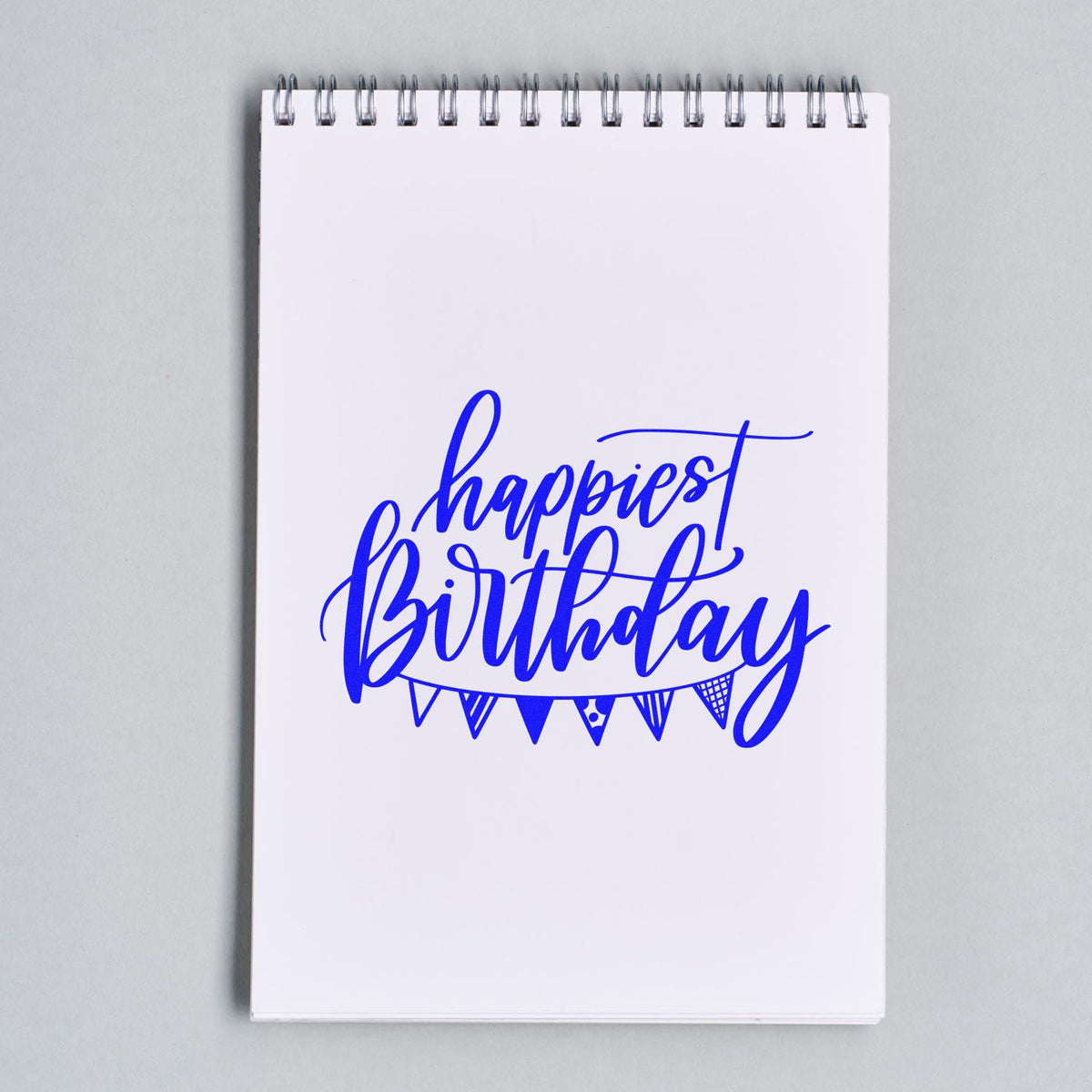 Blue happiest birthday stamp and seal design with bunting on a white spiral notebook.