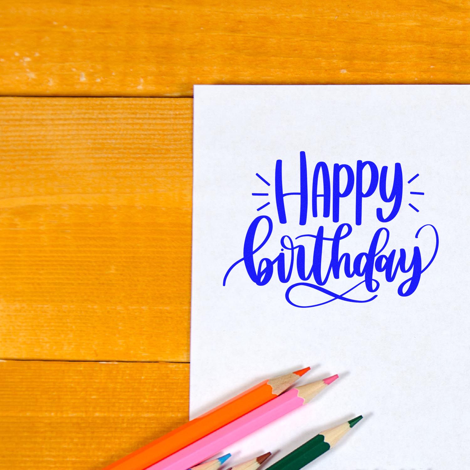 Blue Happy Birthday stamp and seal on white card, with orange, pink, and green pencils on a wooden background