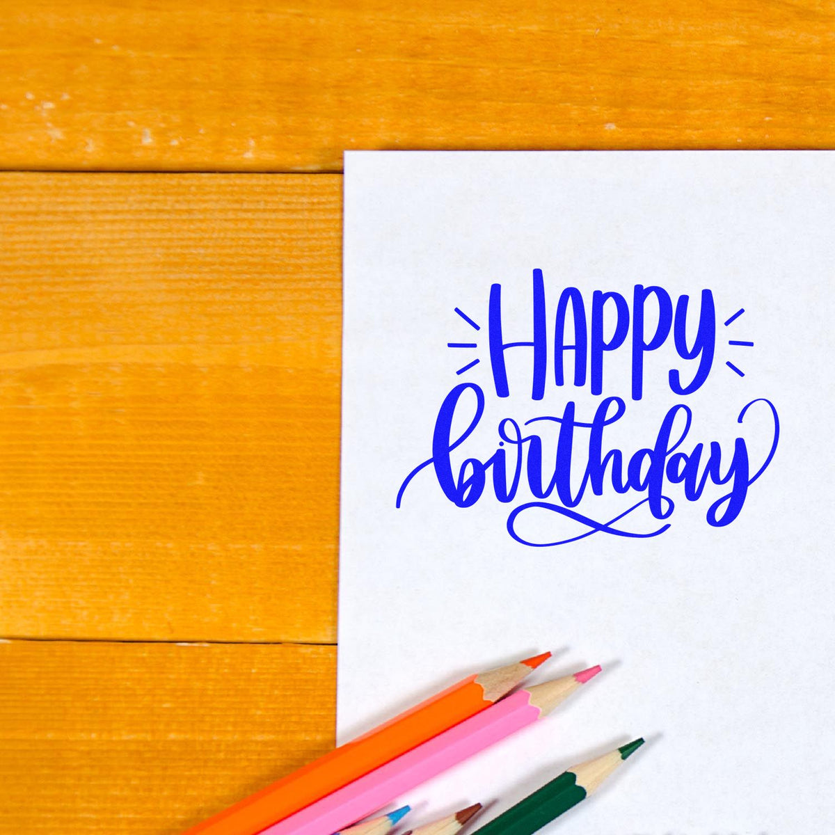 Blue Happy Birthday stamp and seal design on white paper, with pink, orange, and green colored pencils on orange wood.