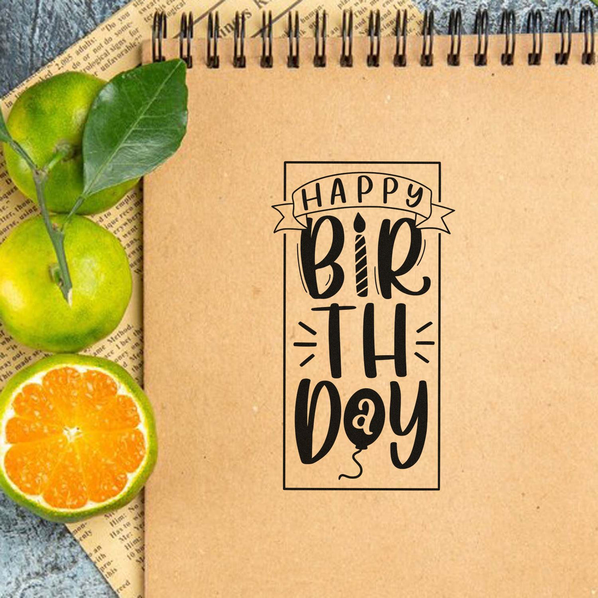 Kraft notebook featuring a black Happy Birthday stamp design, with green oranges, leaves and citrus seal accents.