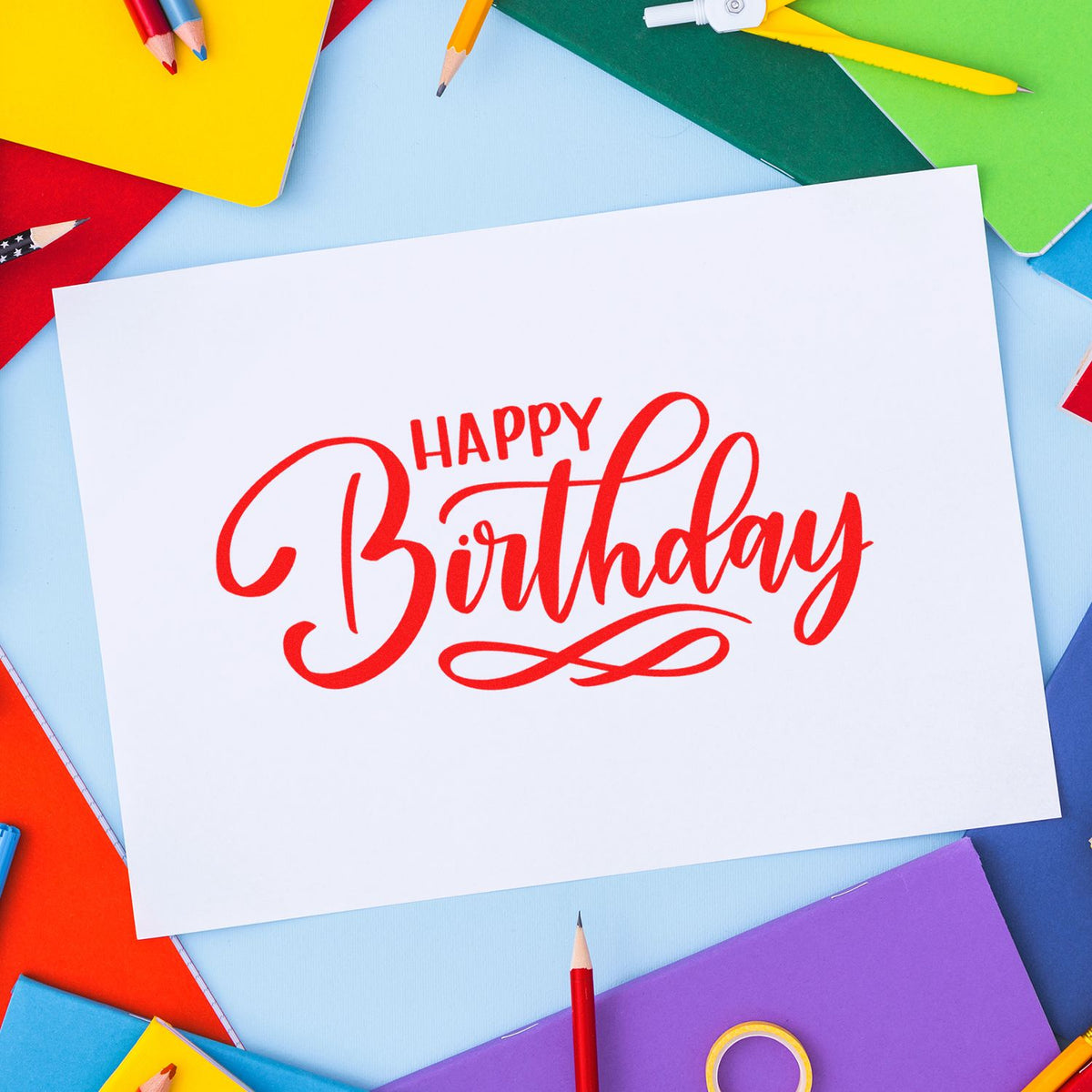 Red Happy Birthday stamp and seal on white card, surrounded by colorful notebooks and pencils.