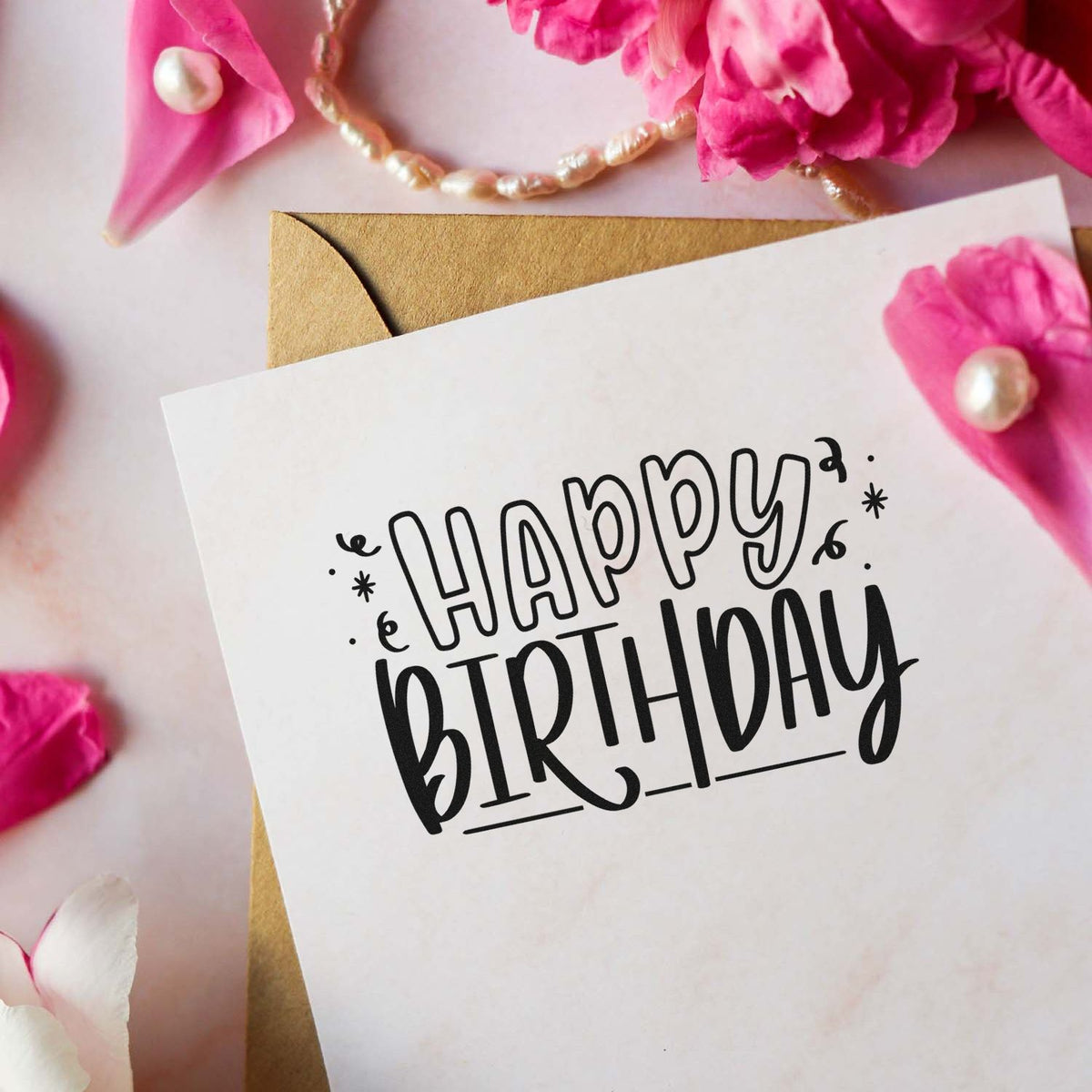 Black Happy Birthday stamp and seal on white card, with gold envelope, pink flowers, and pearl accents.