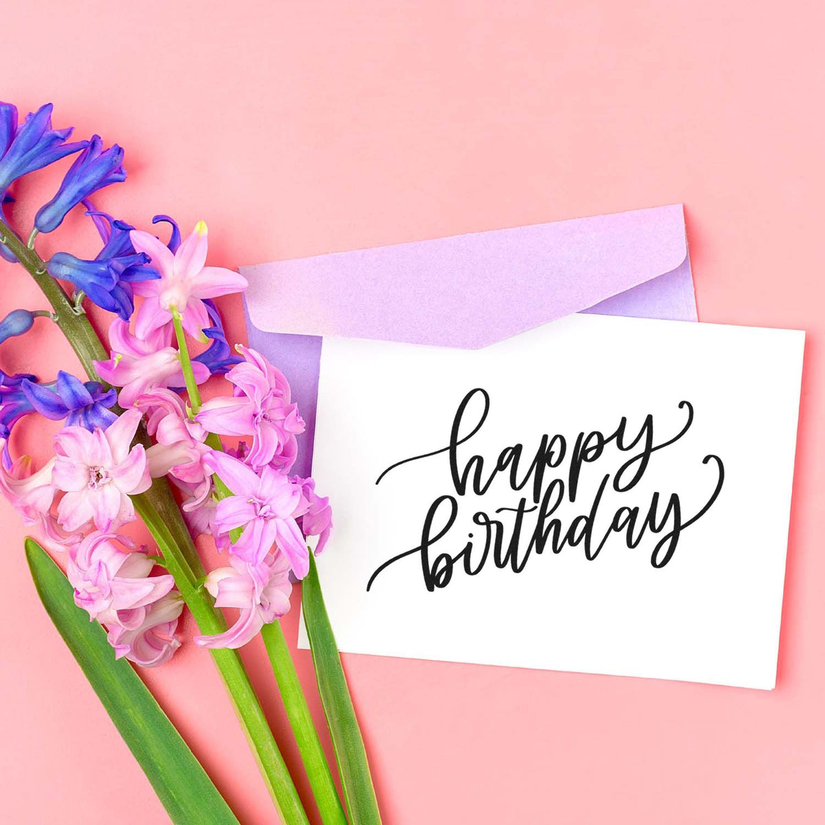 Happy birthday stamp and seal on white card with lavender envelope and pink-purple hyacinths on pastel pink background.