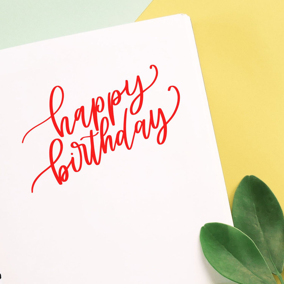 Red happy birthday stamp seal on white card, with pastel yellow and green background and green leaves.
