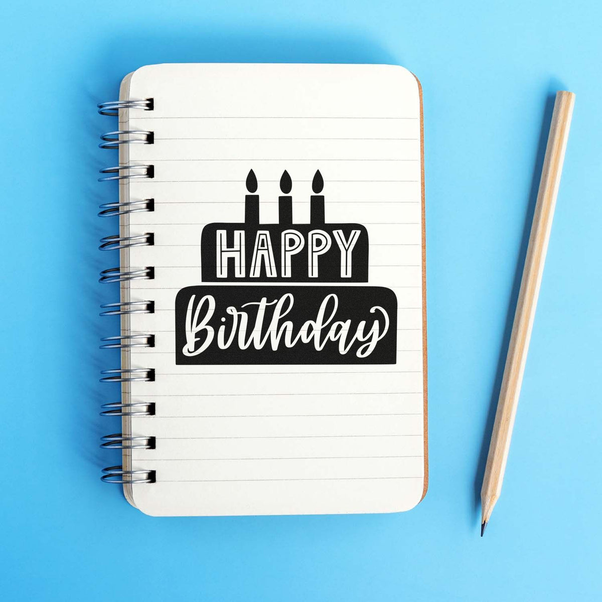 Black Happy Birthday cake stamp on a white spiral notebook, with pencil, on a bright blue background.