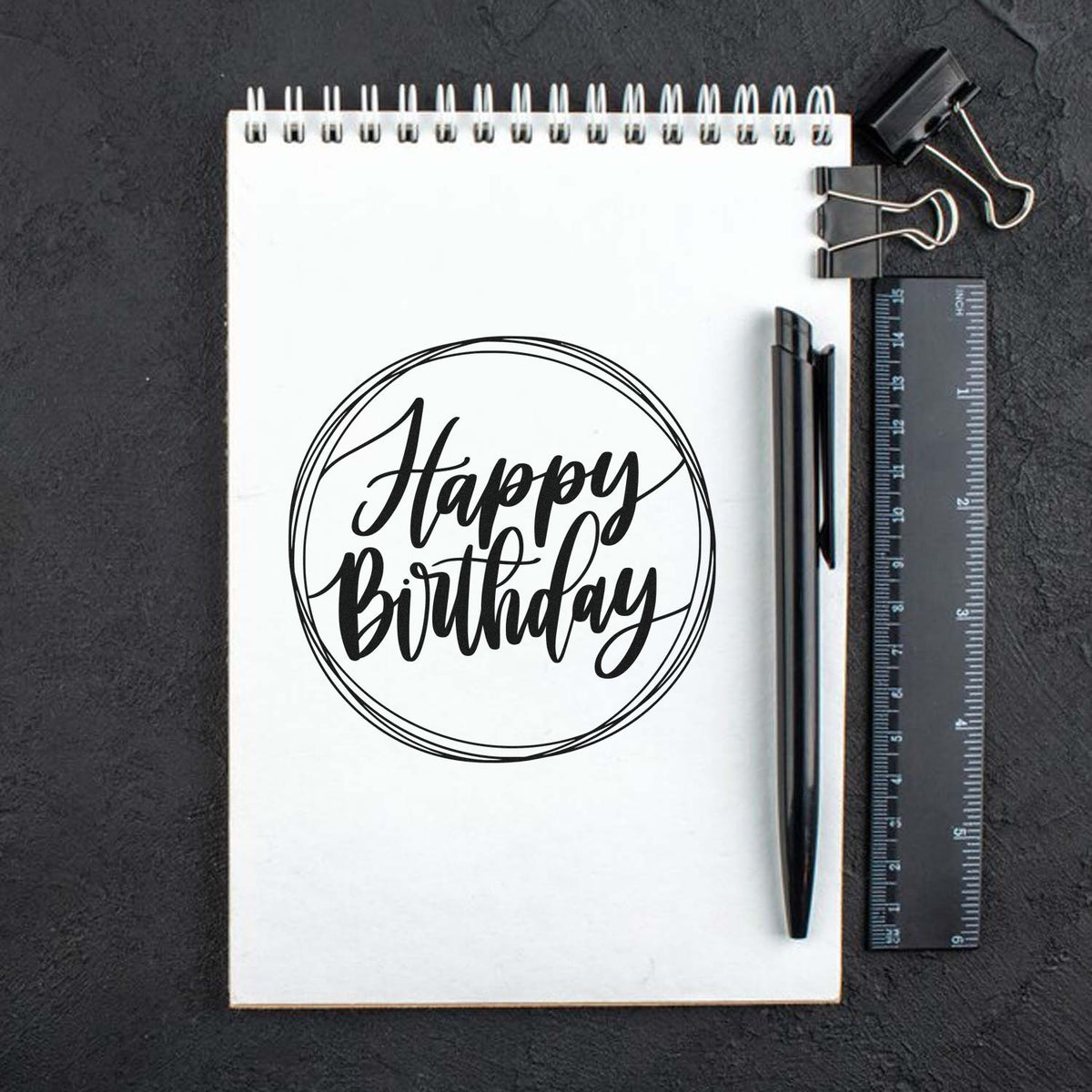 Black Happy Birthday stamp seal on white notepad with pen, ruler, and binder clip on dark background.