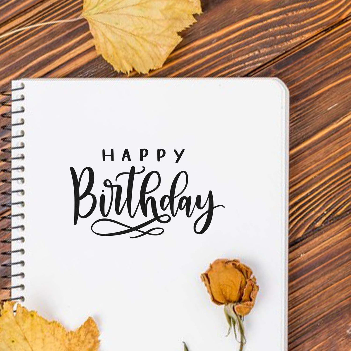 Black Happy Birthday stamp on a white spiral notebook, accented by yellow leaves and a dried flower on rustic wood.
