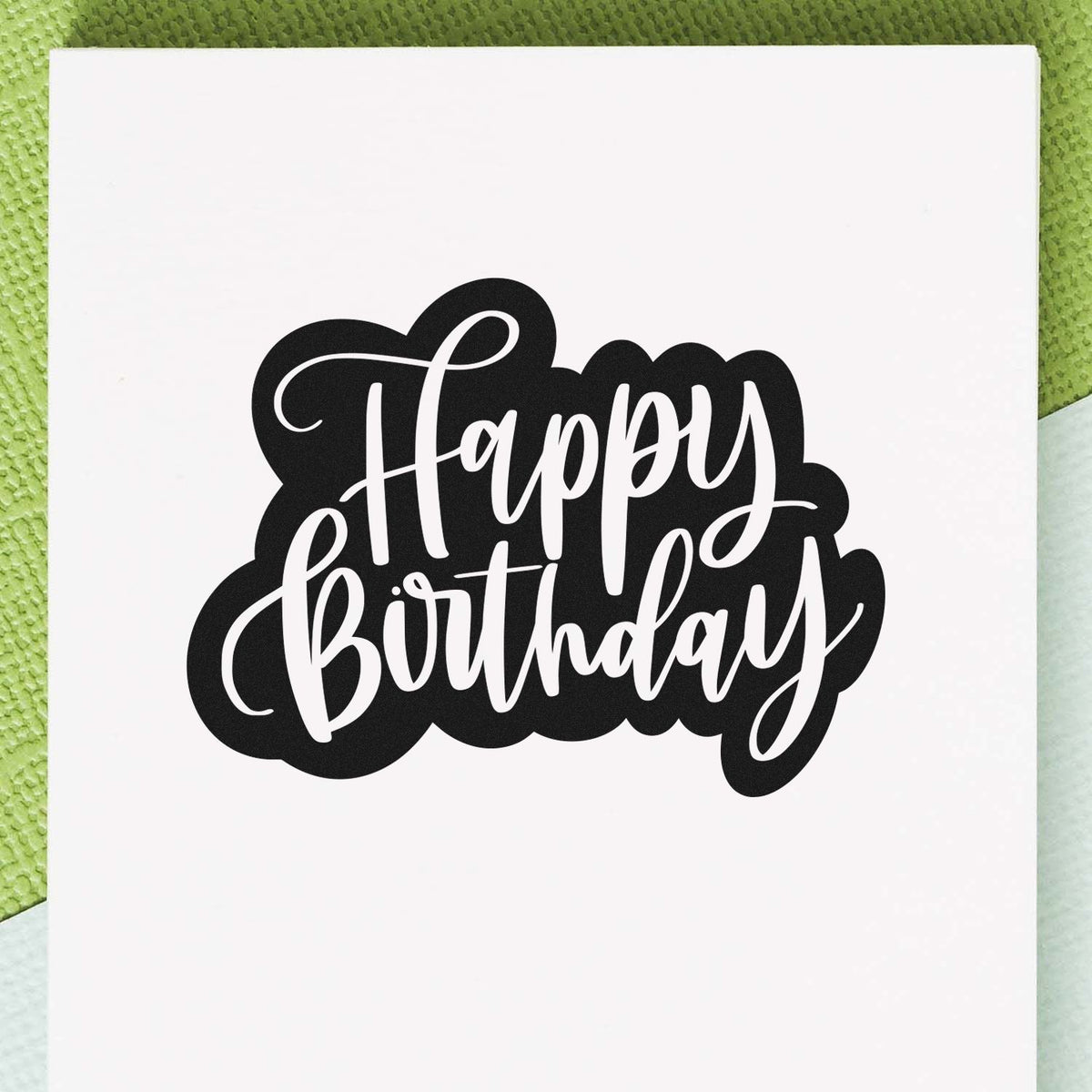 Black Happy Birthday stamp seal with white script on a white card against a textured green background.