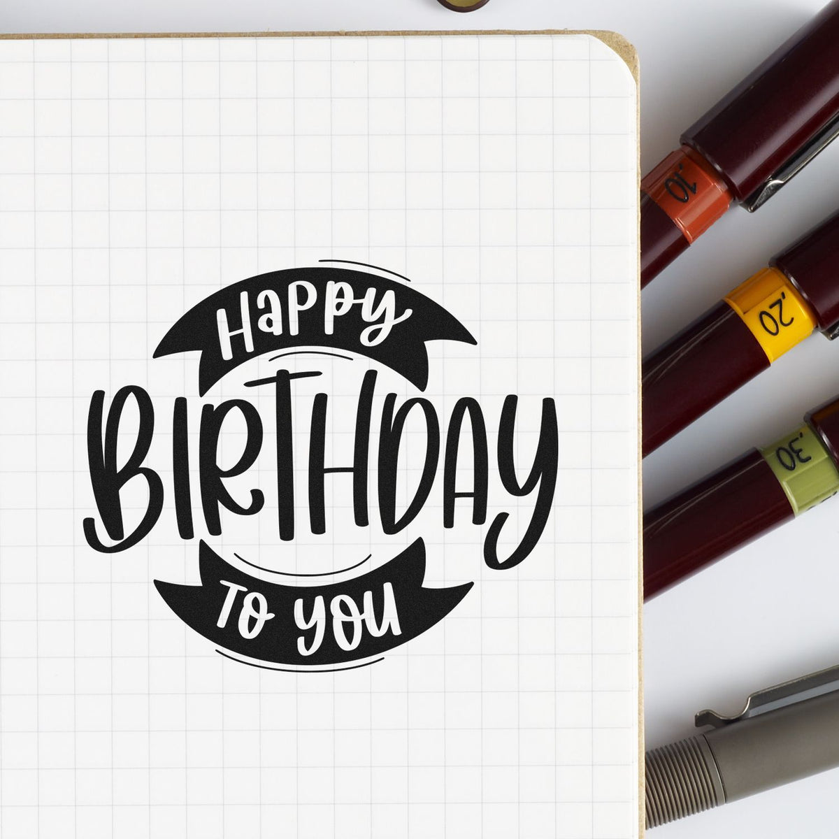 Black Happy Birthday To You stamp on grid notebook, beside burgundy seal markers with orange, yellow, and green date bands.