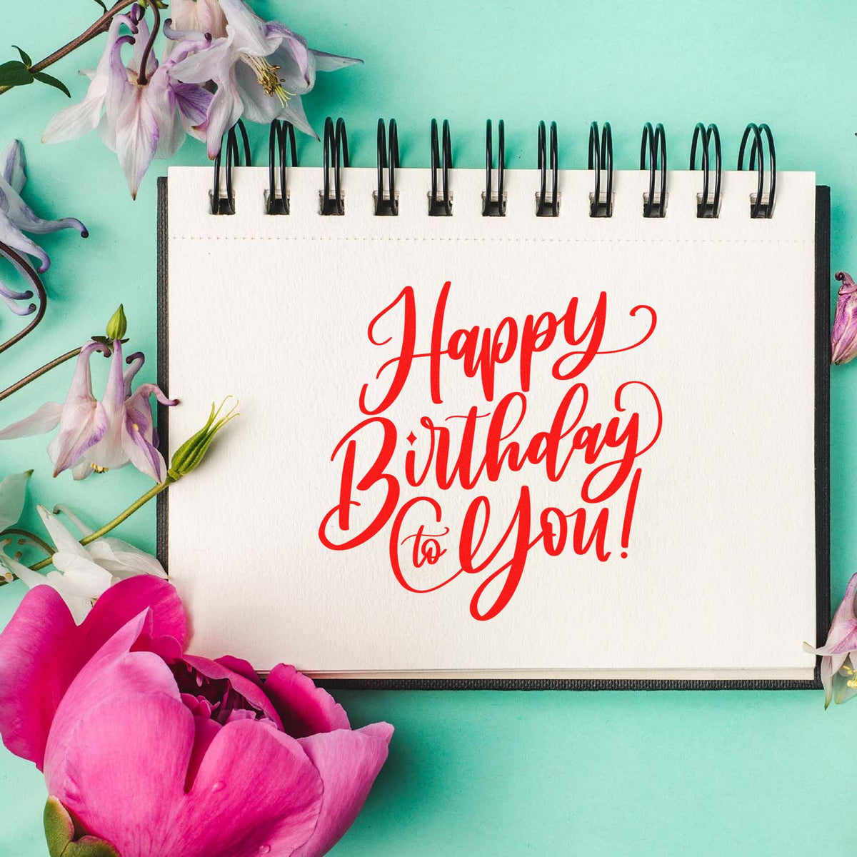 Red Happy Birthday to You! birthday stamp on a white spiral notebook, with pink and purple flowers on a teal background.