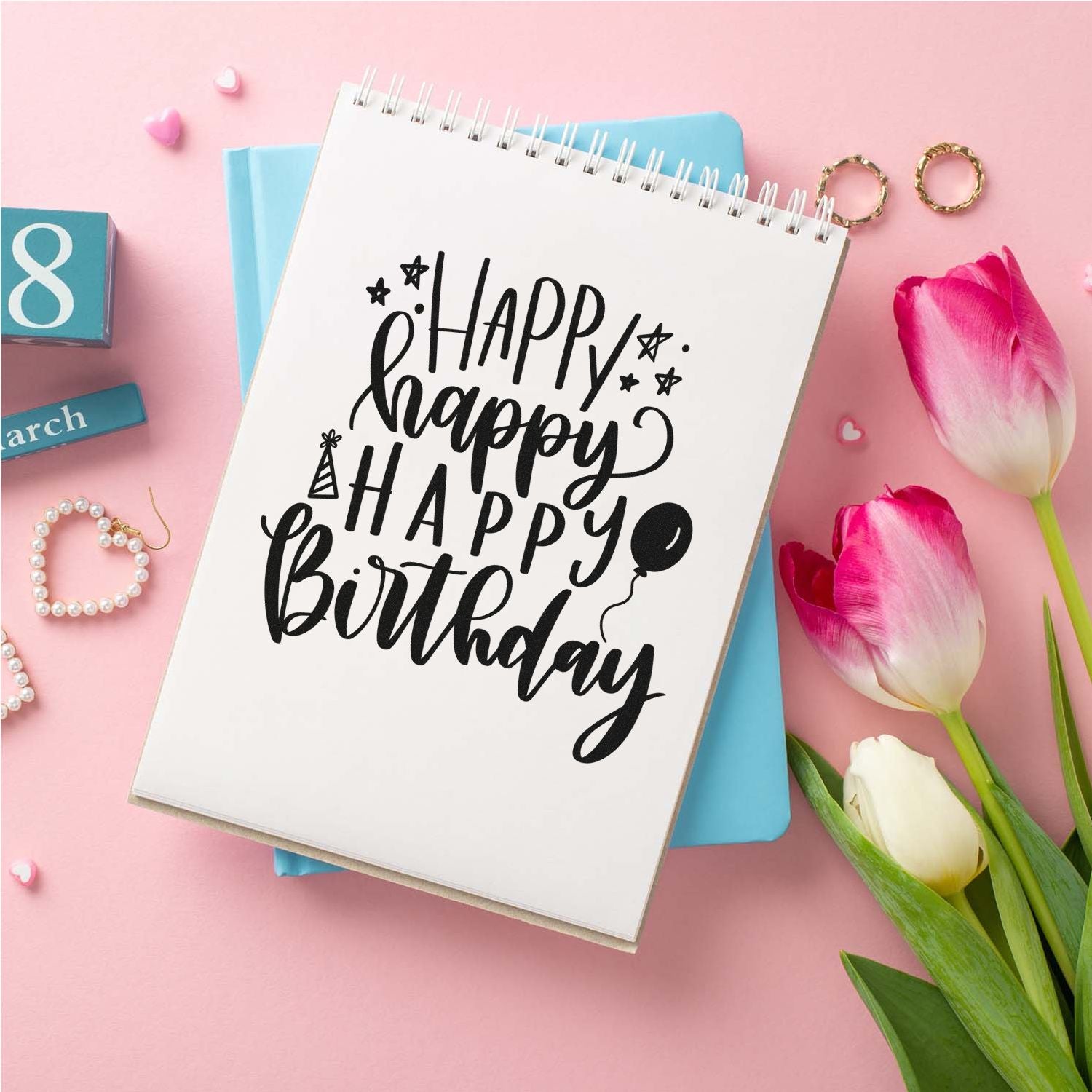 Black Happy Birthday stamp and seal design on white spiral notebook, with pink tulips and pastel accents on a pink background.
