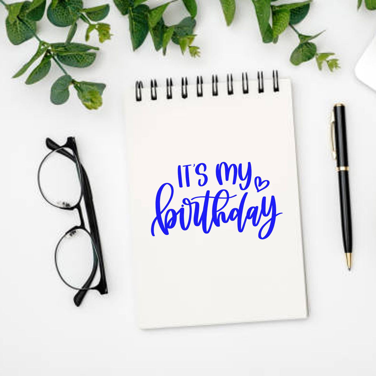 Blue Its my birthday stamp and seal on white notebook, with black glasses, pen, and green leaves.