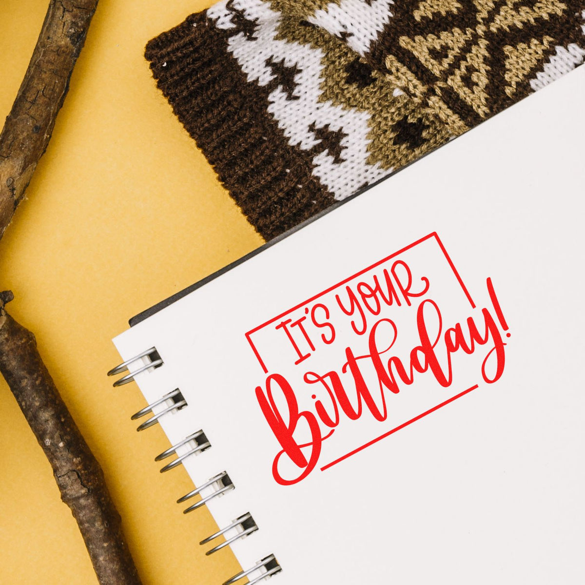 White spiral notebook with red Its your birthday! stamp, on mustard background with brown-and-white knit accents.