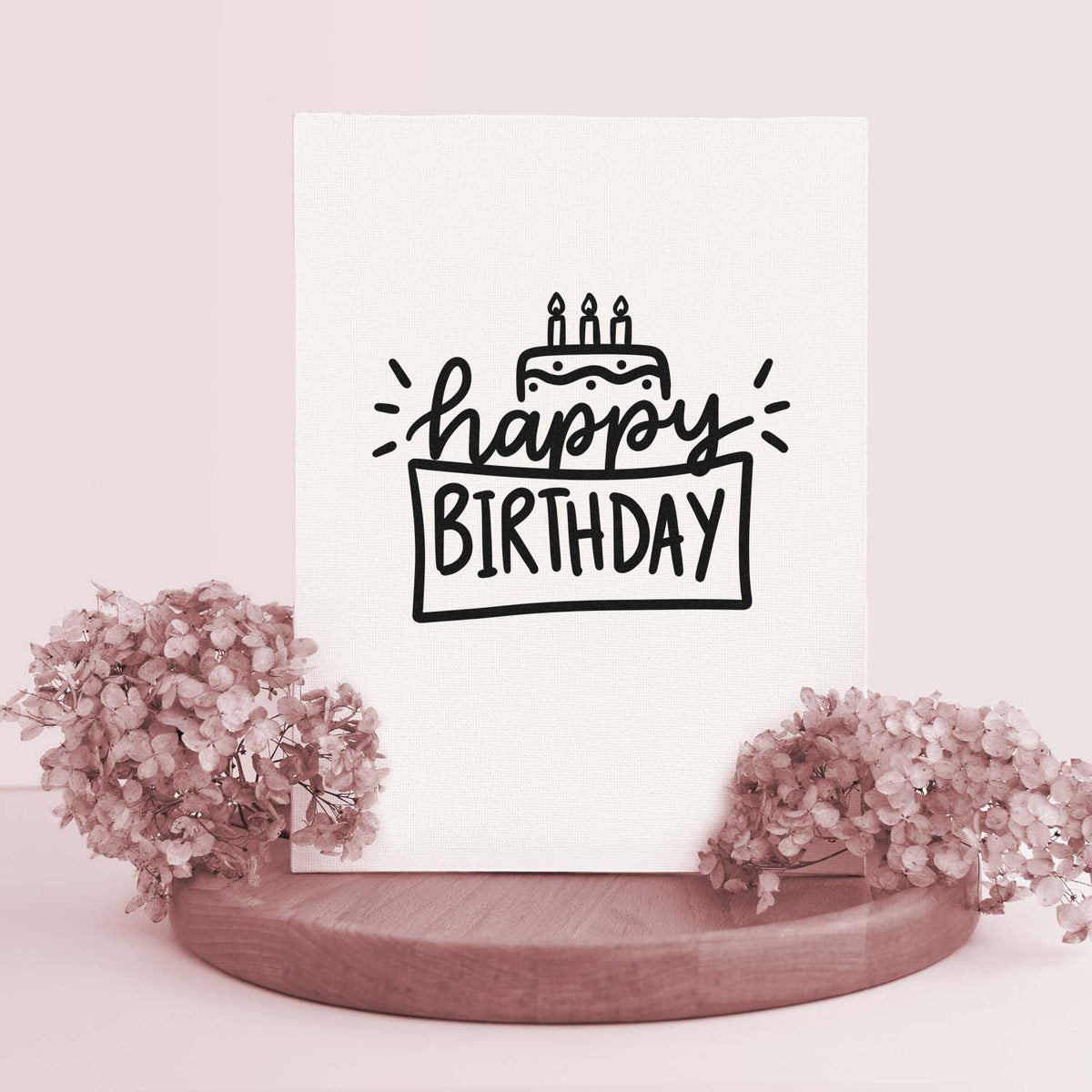Black happy birthday stamp and seal with cake icon on white card, styled with pale pink flowers and a wooden base.