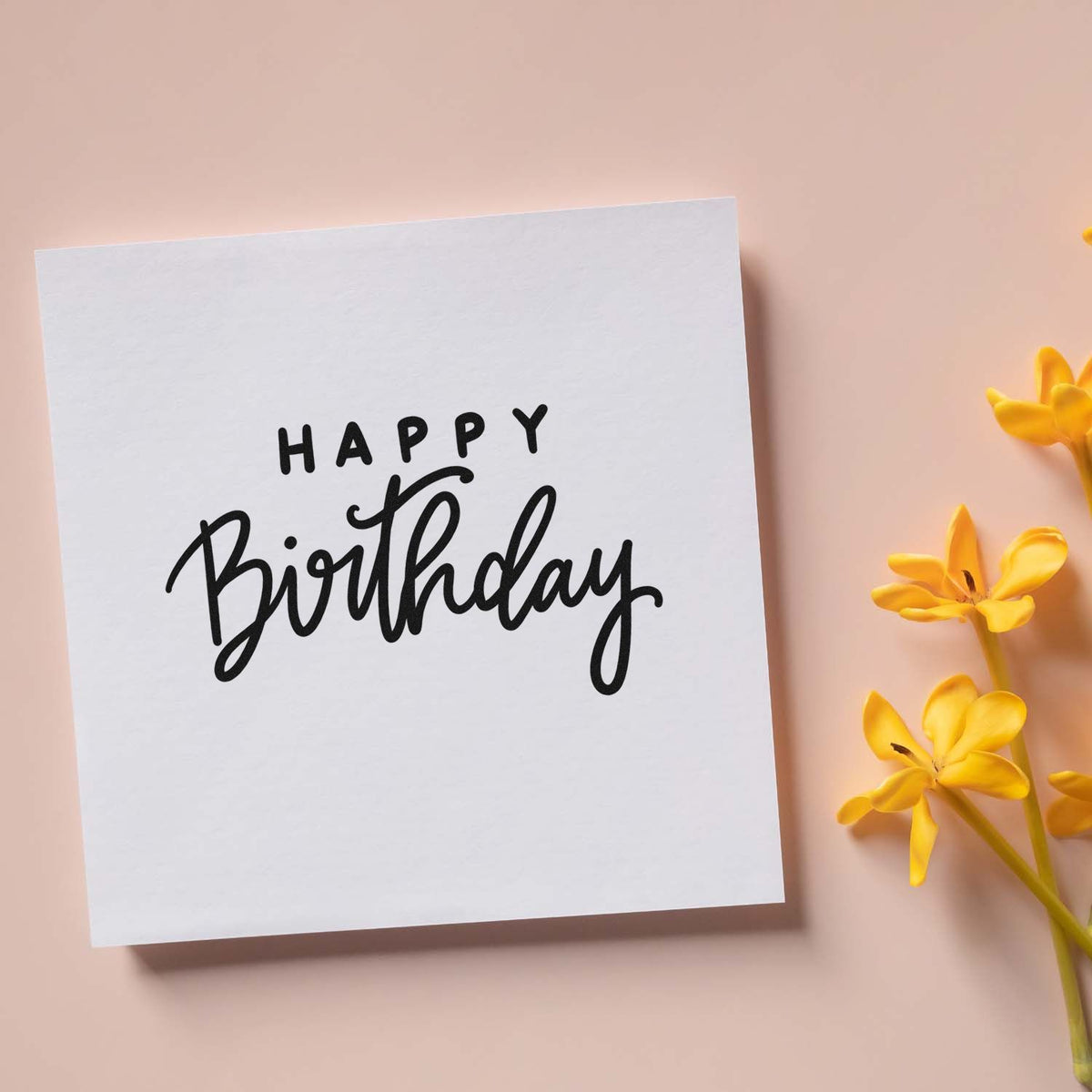 Black Happy Birthday stamp/seal on white card with yellow flowers against a soft peach background.