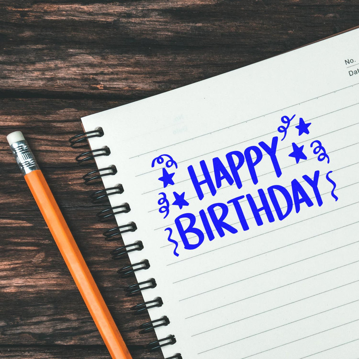 Blue Happy Birthday stamp seal with stars on a white spiral notebook, beside an orange pencil on dark wood.