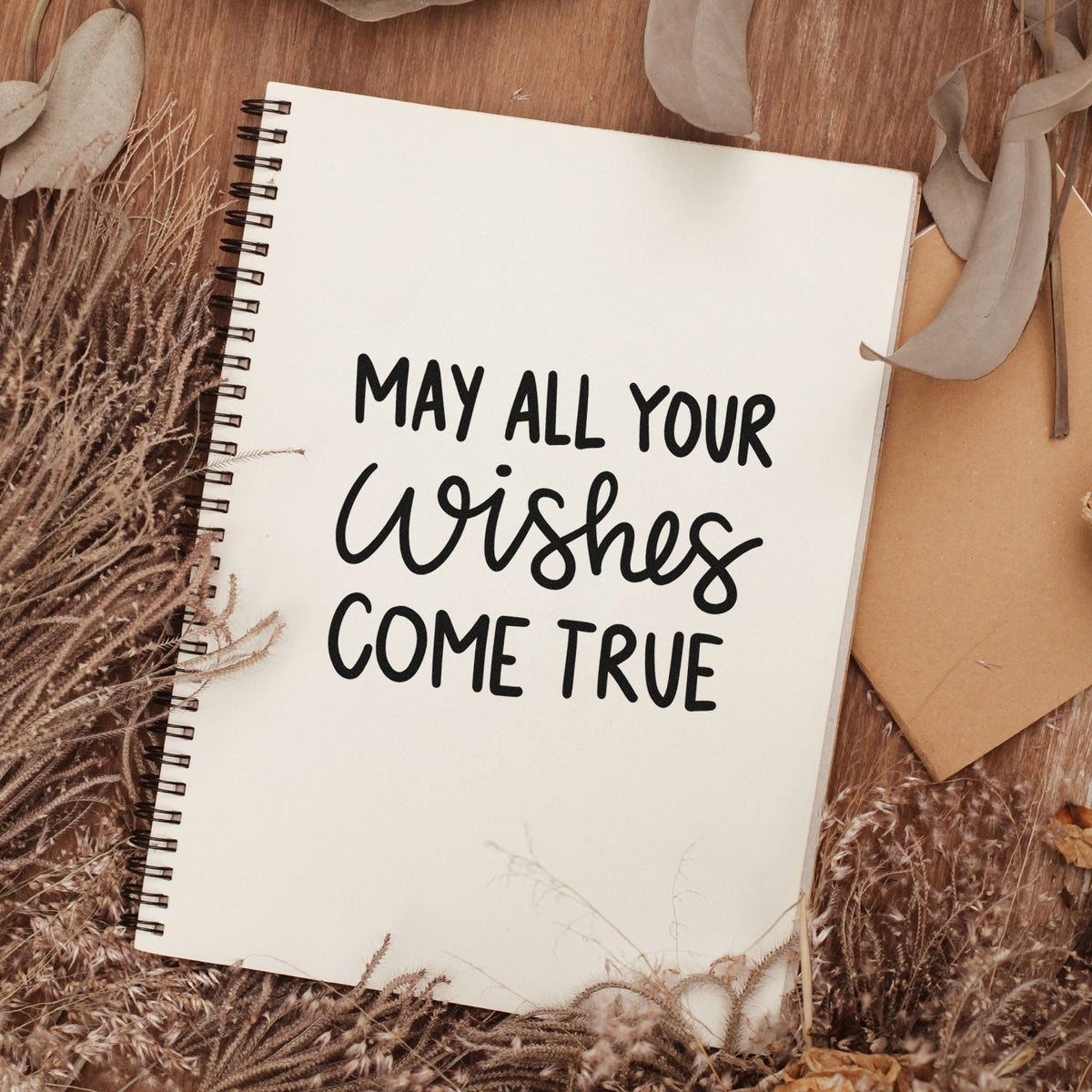 Custom stamp and seal design on cream spiral notebook, with black May All Your Wishes Come True text and dried beige florals.