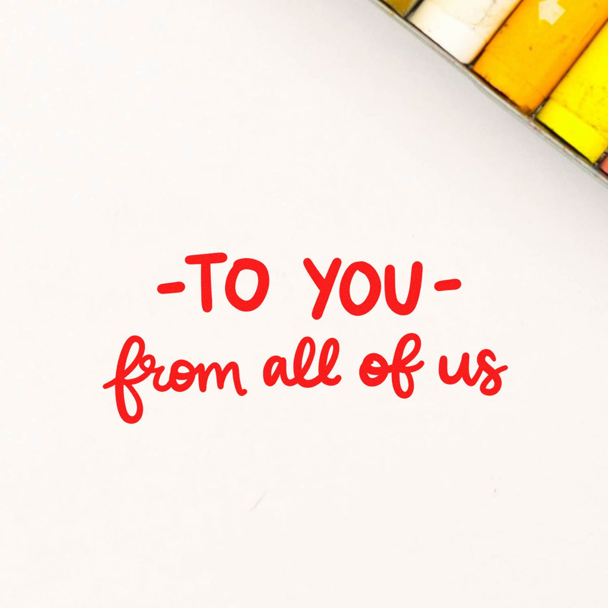 Red rubber stamp seal with handwritten text To you from all of us on a white background with yellow corner accents.
