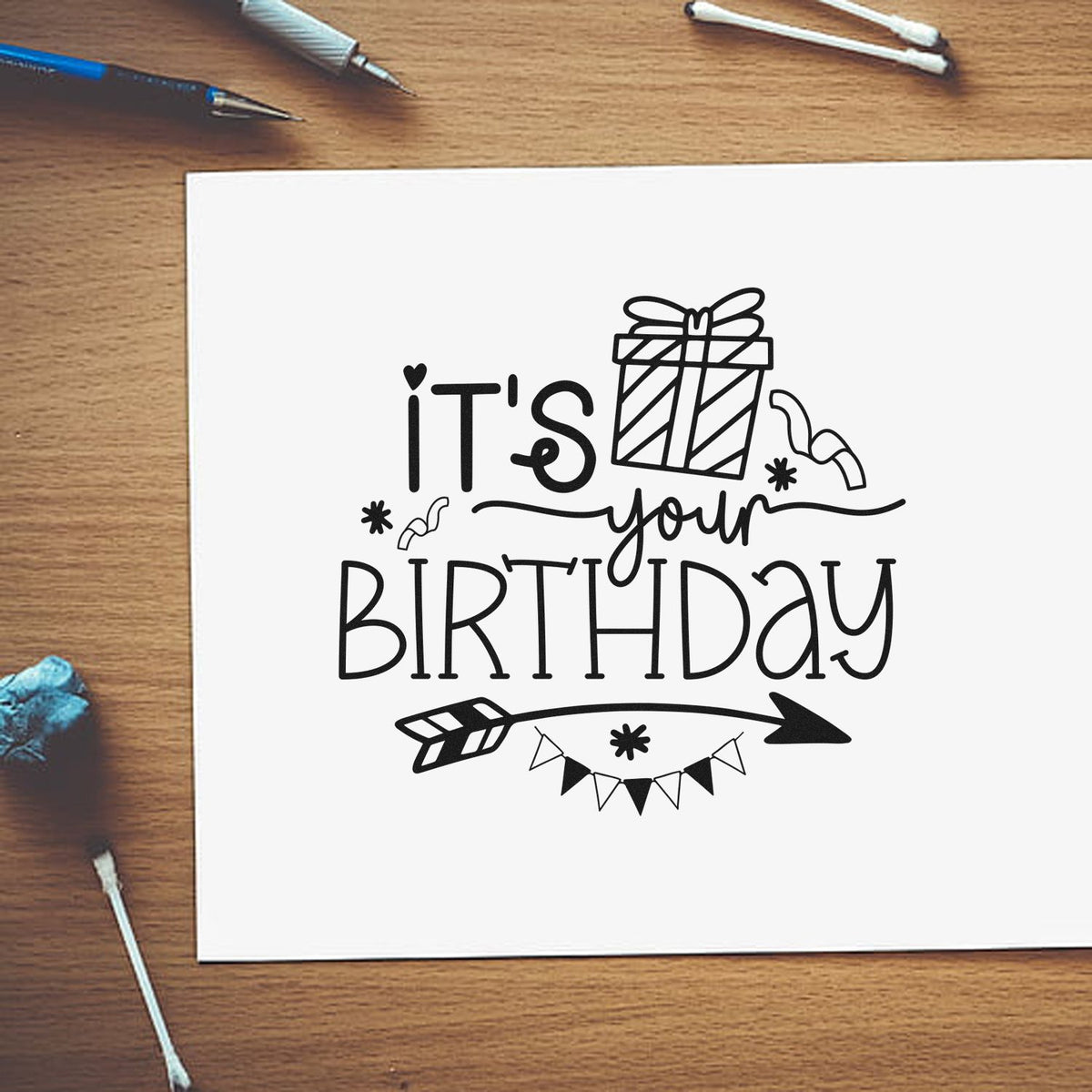 Black birthday stamp and seal design on white paper, with gift icon, arrow, bunting, and craft tools on a wood desk.