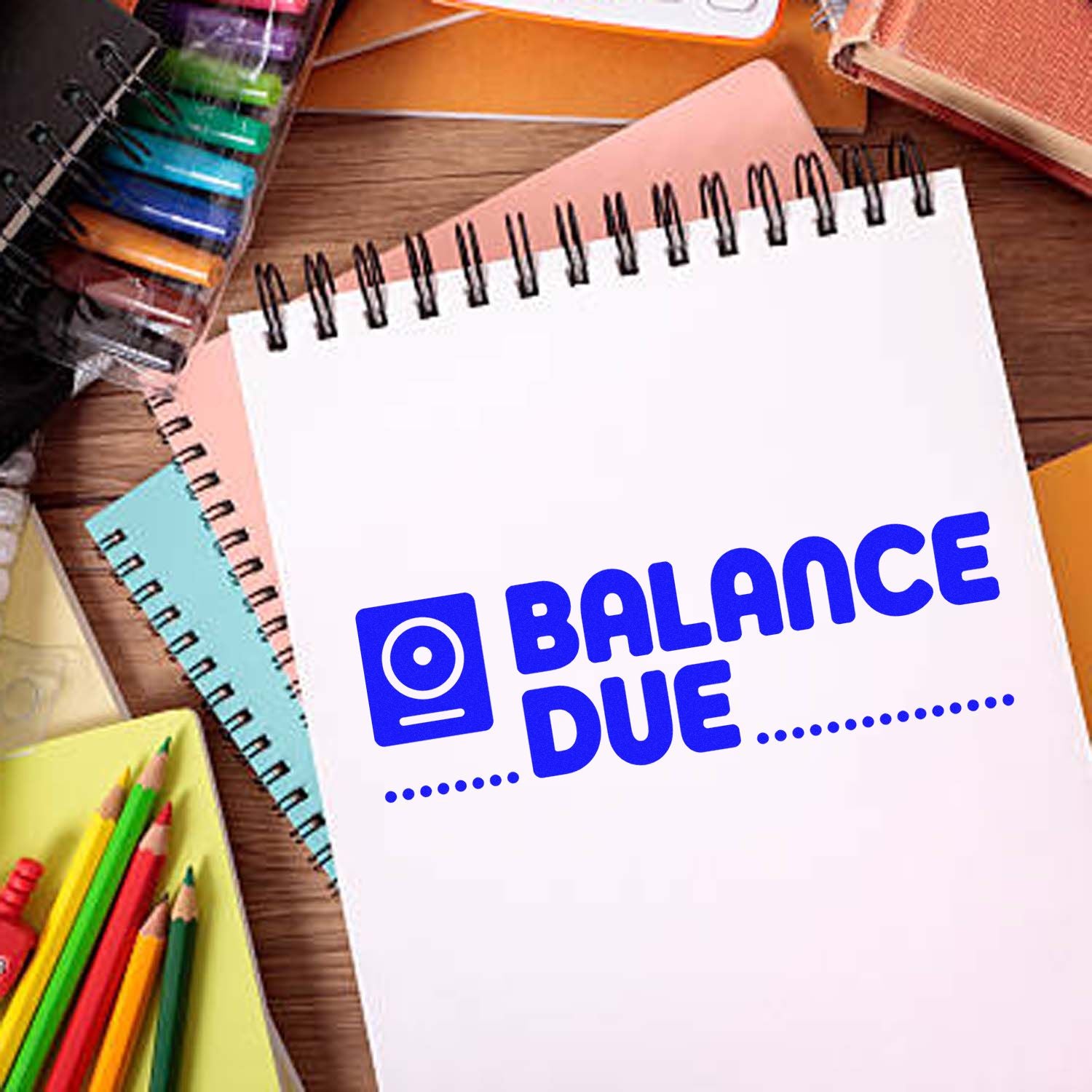 Blue Balance Due stamp design on white spiral notebook, surrounded by colorful stationery on a wooden desk.